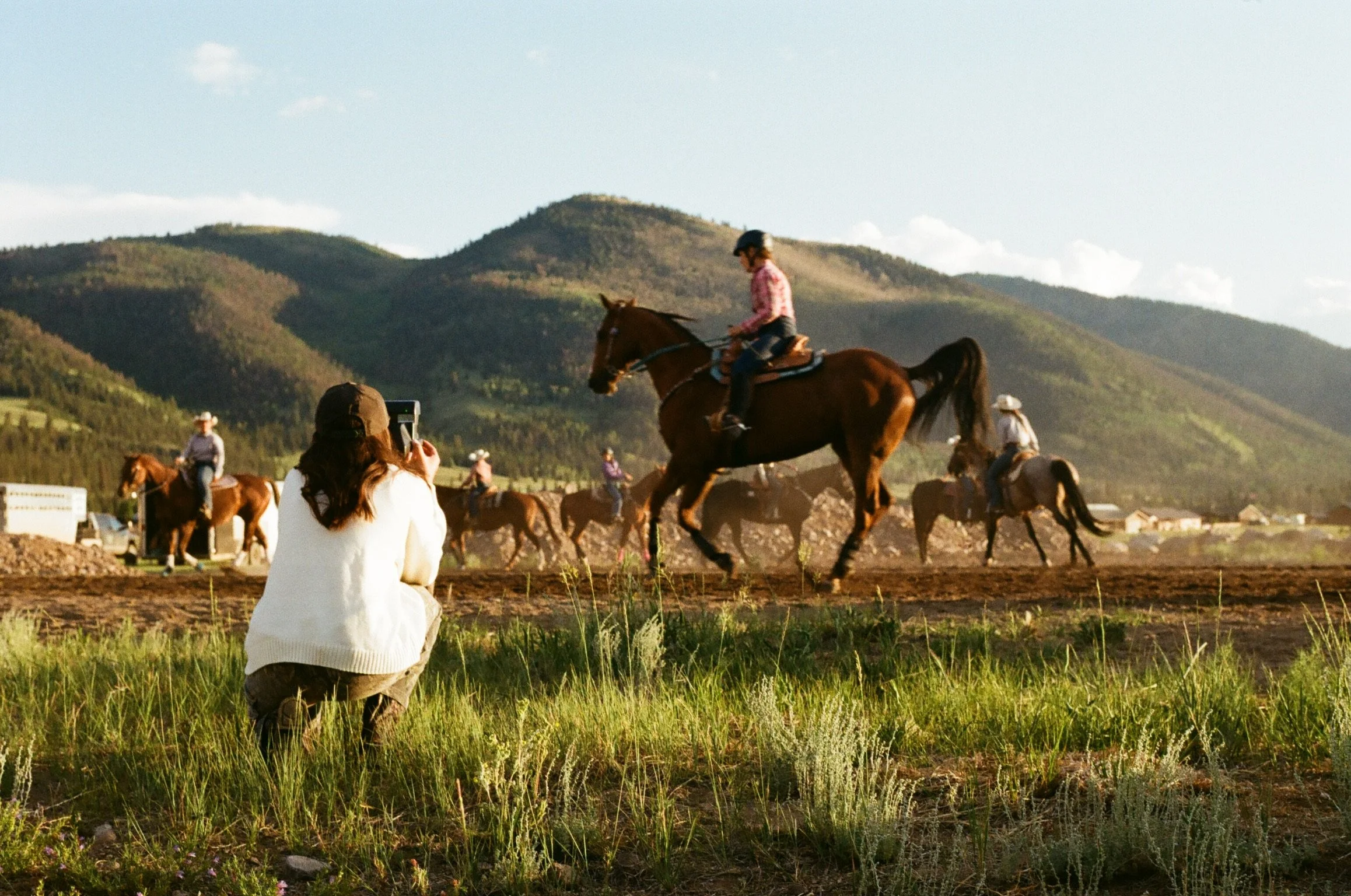 A woman crouching in a grassy field takes a photo of children riding horses in a dirt arena with mountains in the background during the day.