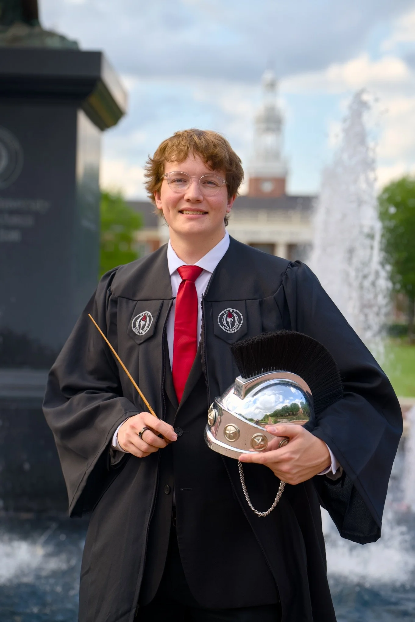 Young man in graduation gown and red tie holding a silver trophy wearing glasses, outdoors with fountain, building, and cloudy sky in background.