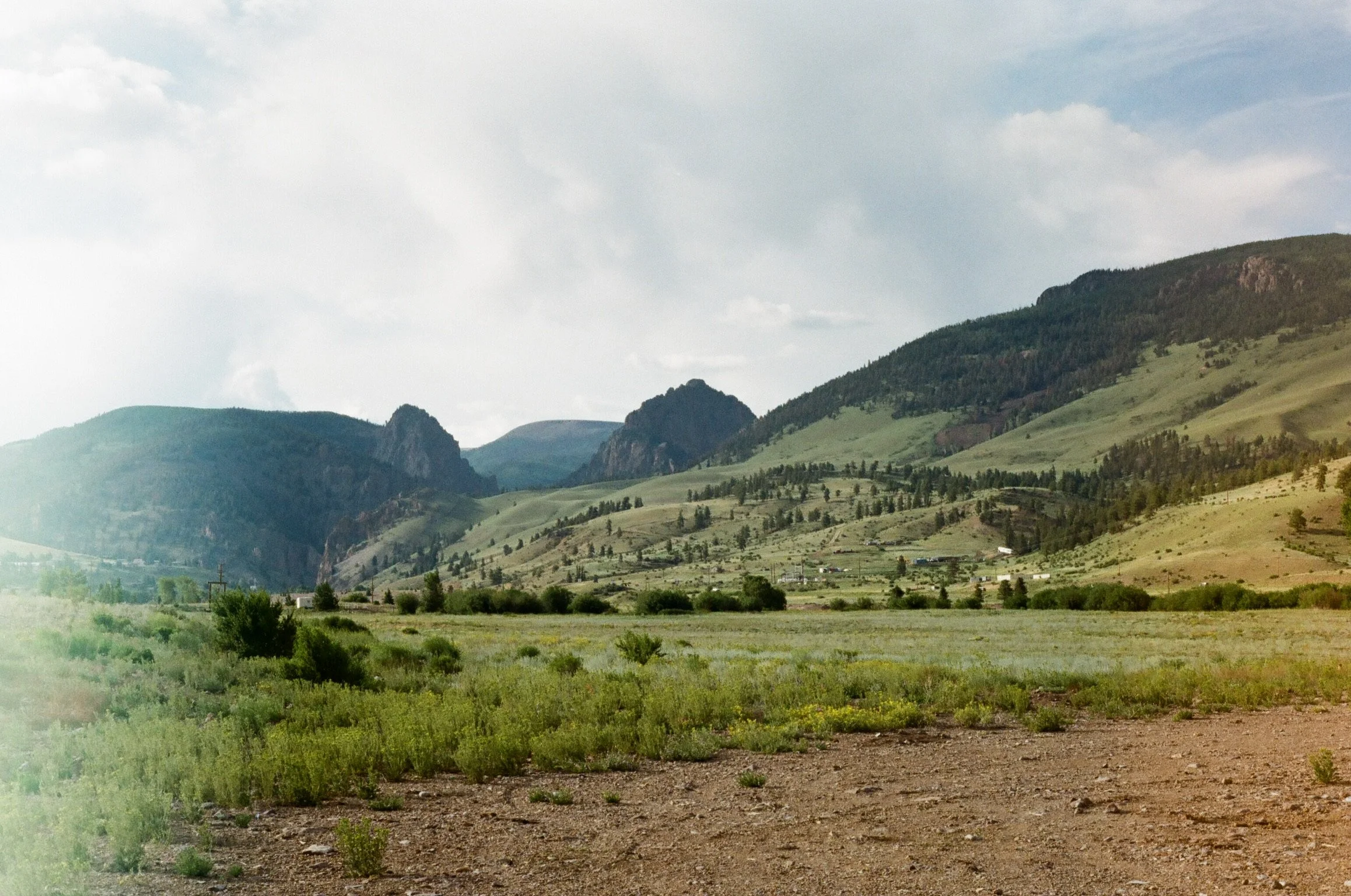 Scenic view of green mountainous landscape with rolling hills, sparse trees, and distant rocky formations under a cloudy sky.