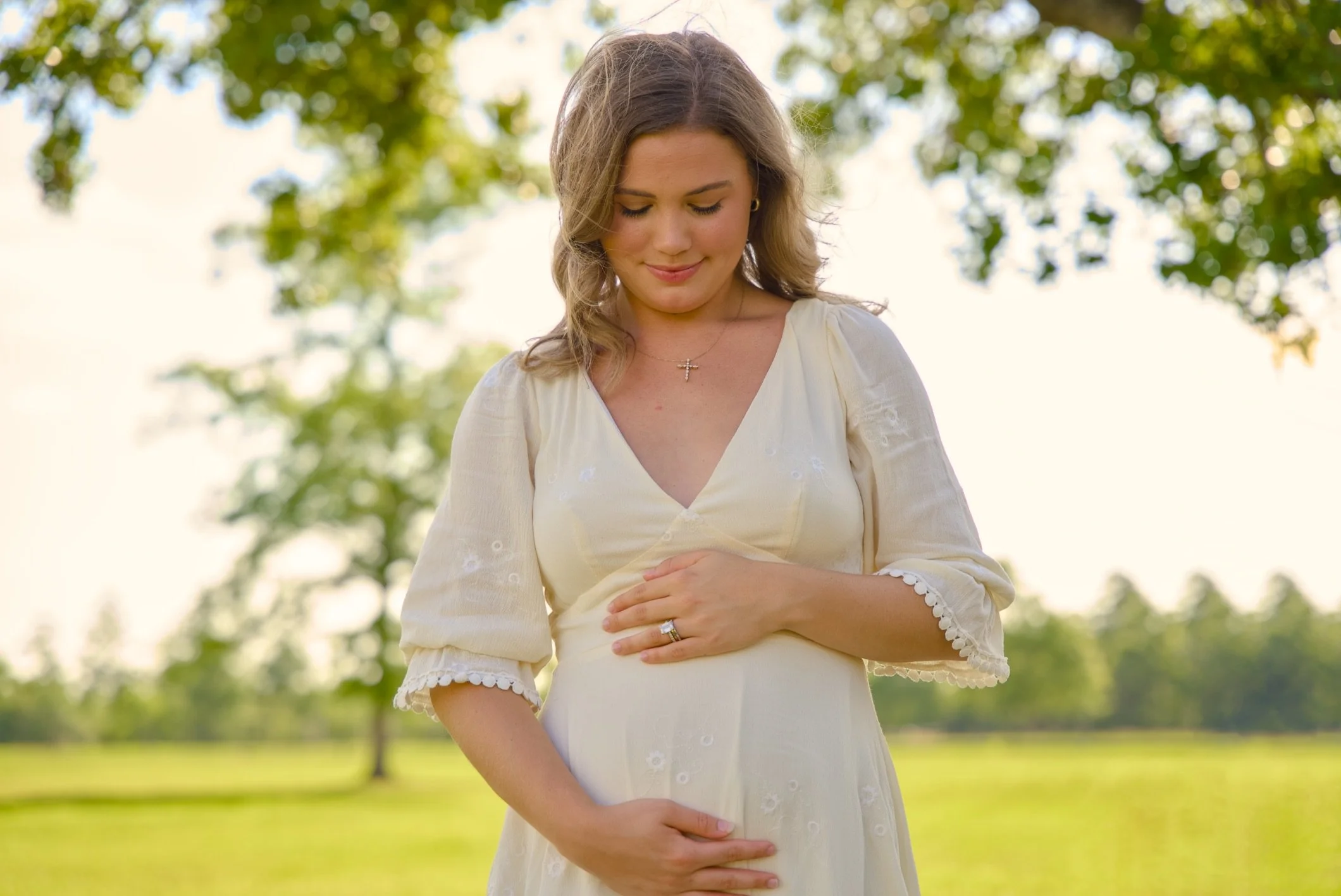 A pregnant woman with light brown hair in a half-up style, wearing a light yellow dress with white embroidery, stands outdoors in a park with green trees and grass, smiling gently with her eyes closed, holding her belly with both hands.