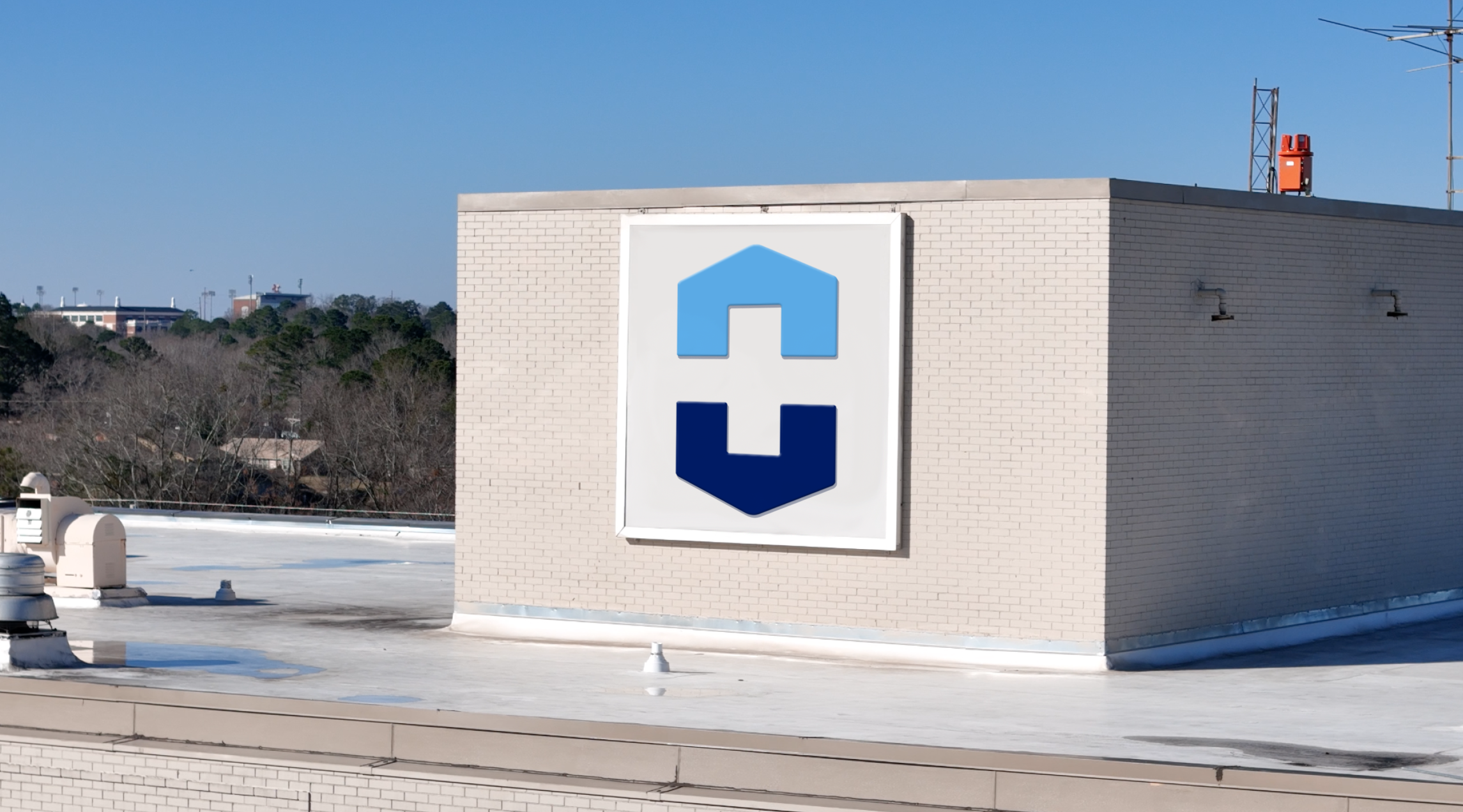 Rooftop view of a building with a large sign featuring a blue house icon and a stylized blue letter H beneath it, with a backdrop of trees and a blue sky.