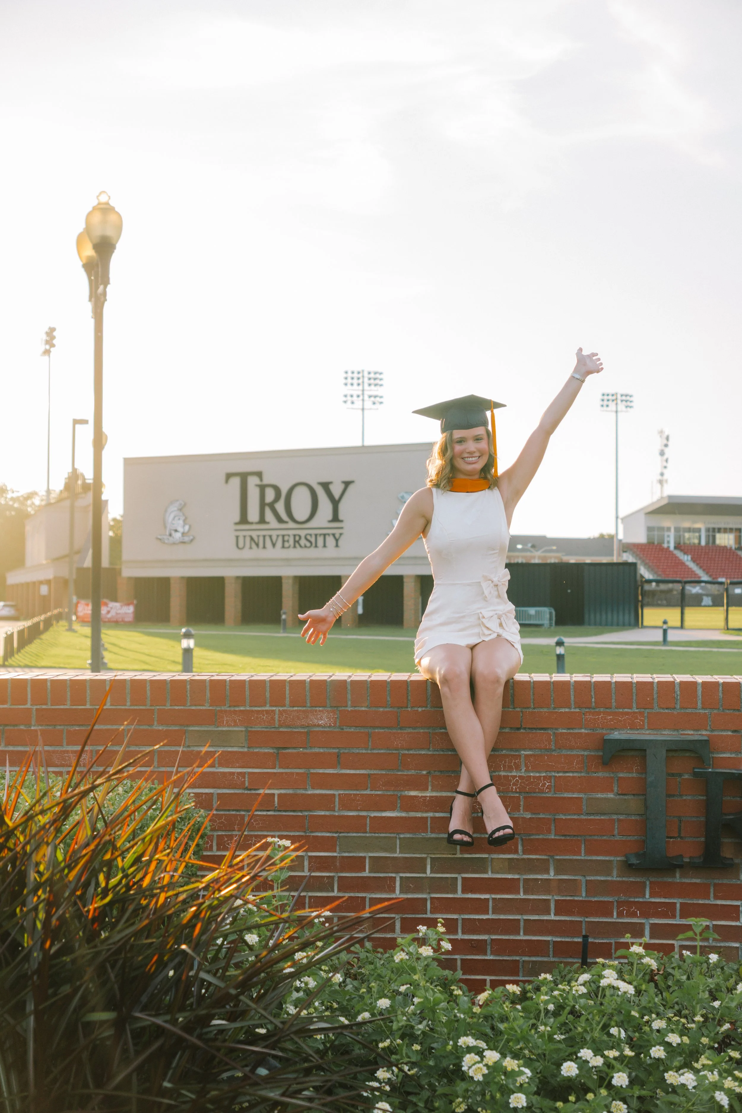 A young woman in a white dress and black heels sitting on a brick wall with one leg crossed over the other, smiling and raising her arms in celebration, wearing a graduation cap, in front of a sign reading 'Troy University' on a college campus.