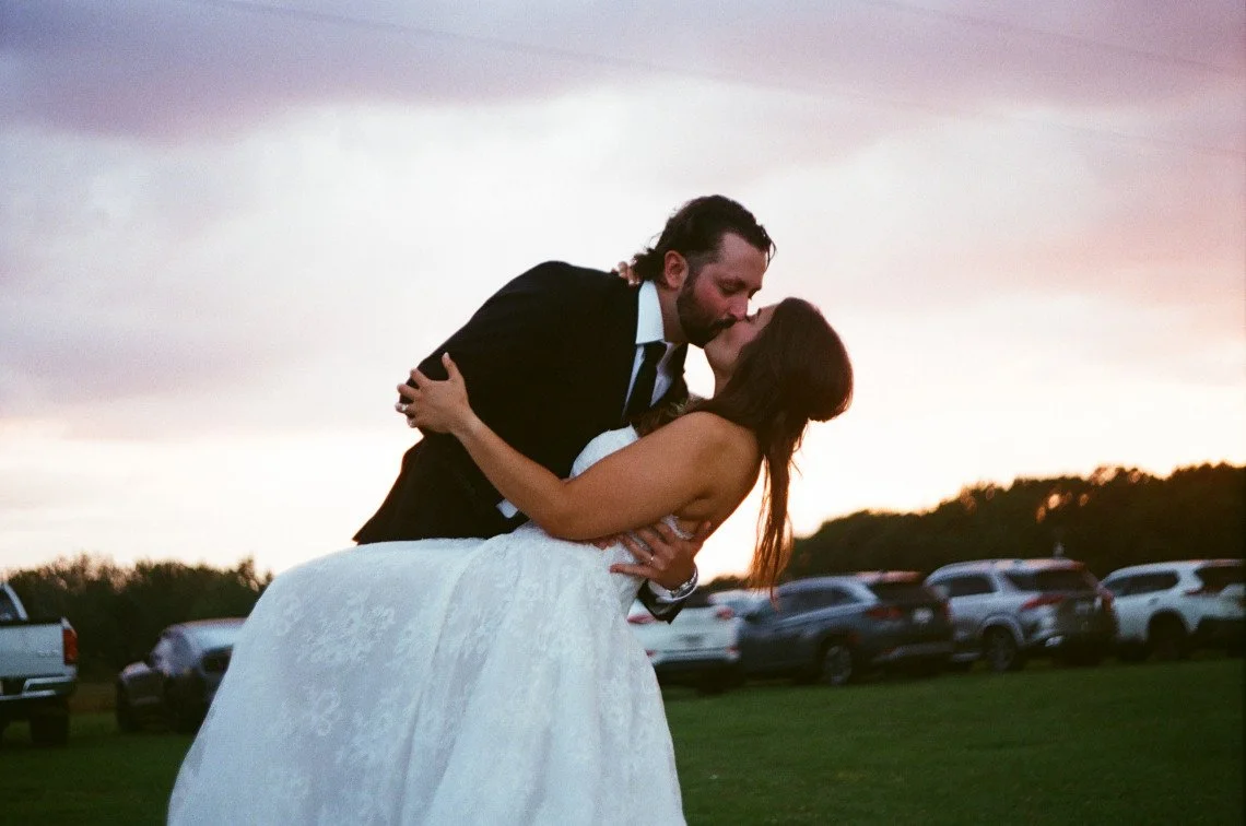 A couple in wedding attire sharing a kiss outdoors at sunset, with parked cars in the background.