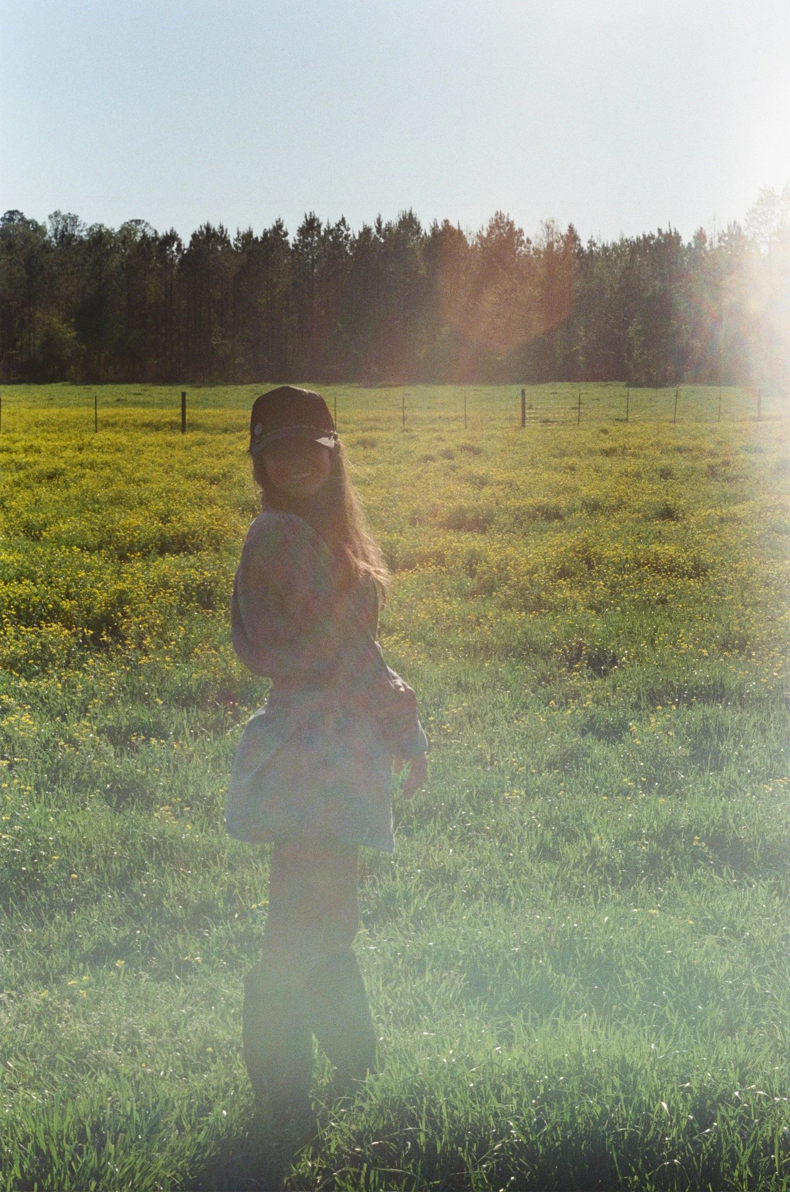 A girl wearing a black hat and glasses standing in a grassy field with yellow flowers, trees in the background, and the sun shining from the right side of the image.