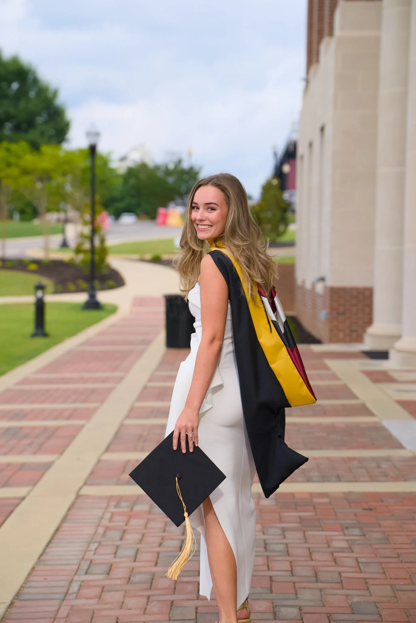 A young woman in a white dress smiling while holding a graduation cap with a gold tassel, standing outdoors on a brick pathway with greenery and buildings in the background.