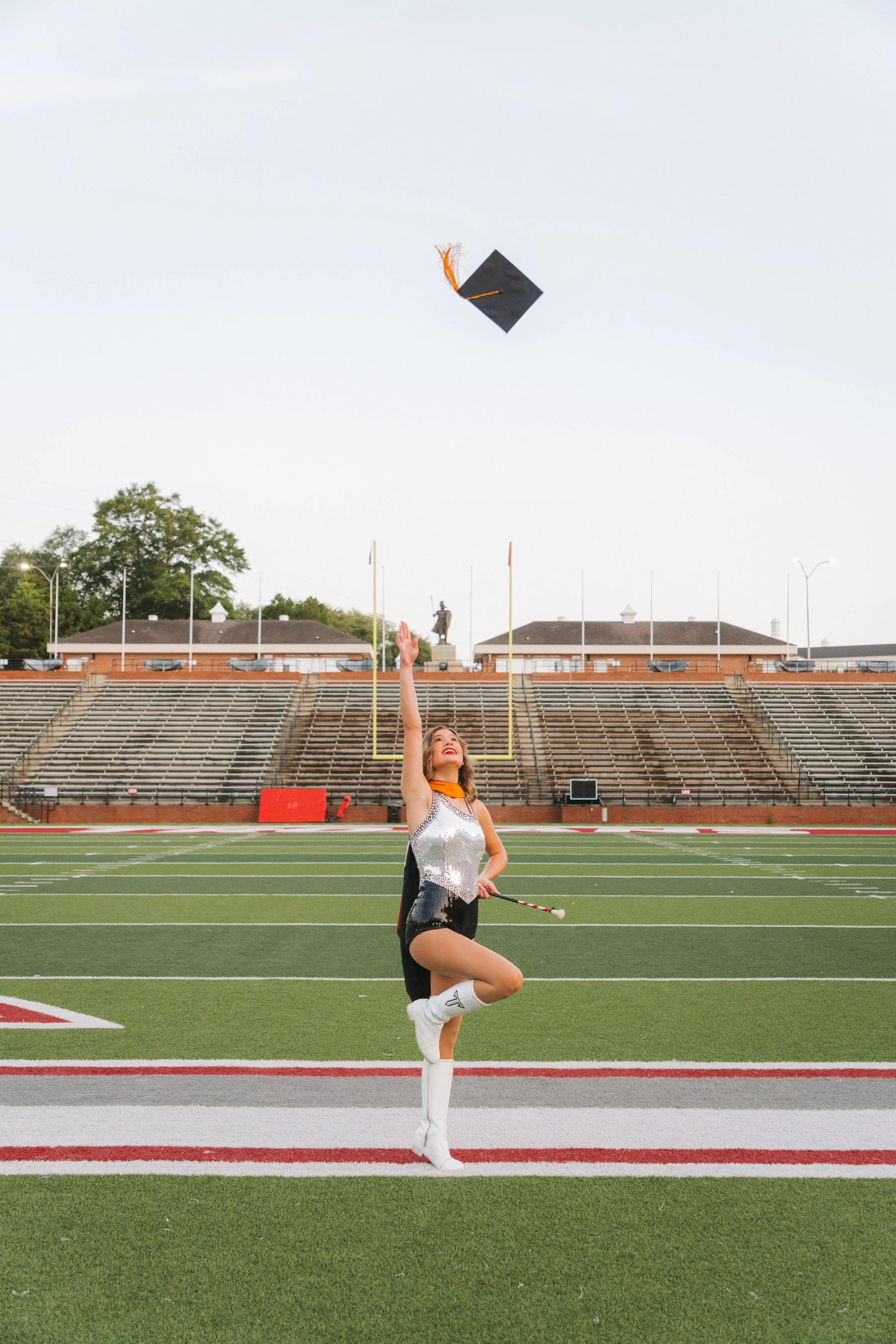 A cheerleader is on a football field, tossing a graduation cap in the air. She is dressed in a shiny silver and black cheerleading uniform with white sneakers, and is standing on one leg with her arms raised, celebrating. The background shows stadium