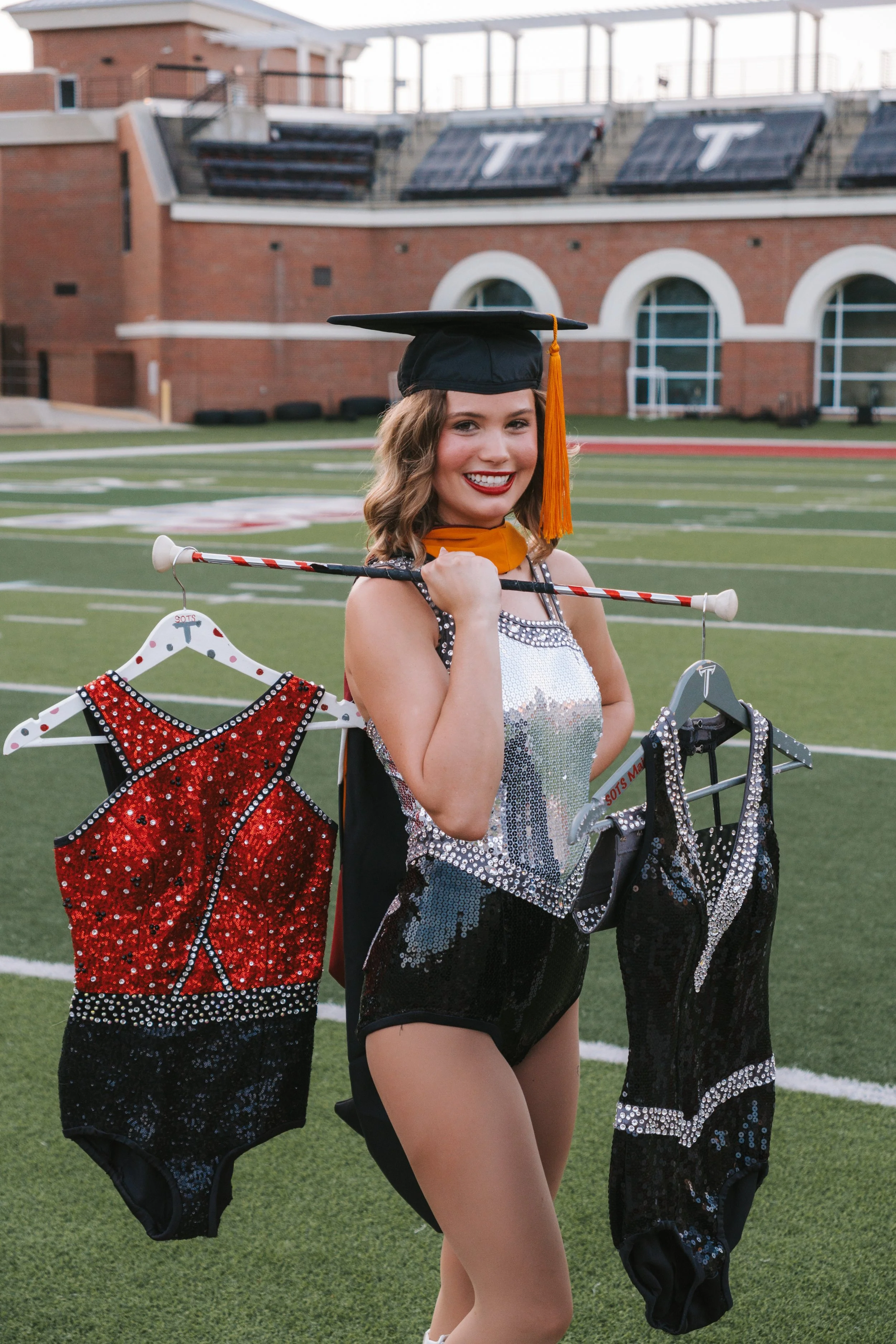 Young woman in a graduation cap and gown, holding baton, standing on a football field with cheerleading uniforms and hootchie on display.