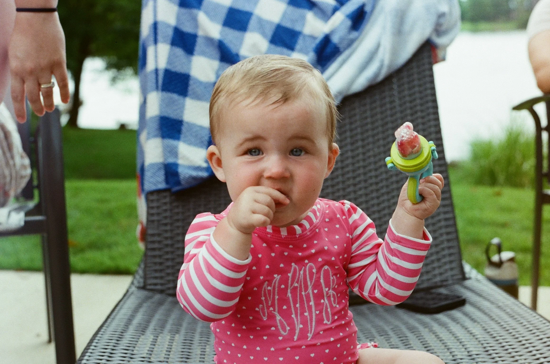 A young girl with blonde hair wearing a pink and white striped shirt holding a toy sword with a red, white, and green handle. She is sitting on a black outdoor chair outdoors with a man wearing a blue and white checkered shirt in the background. Ther