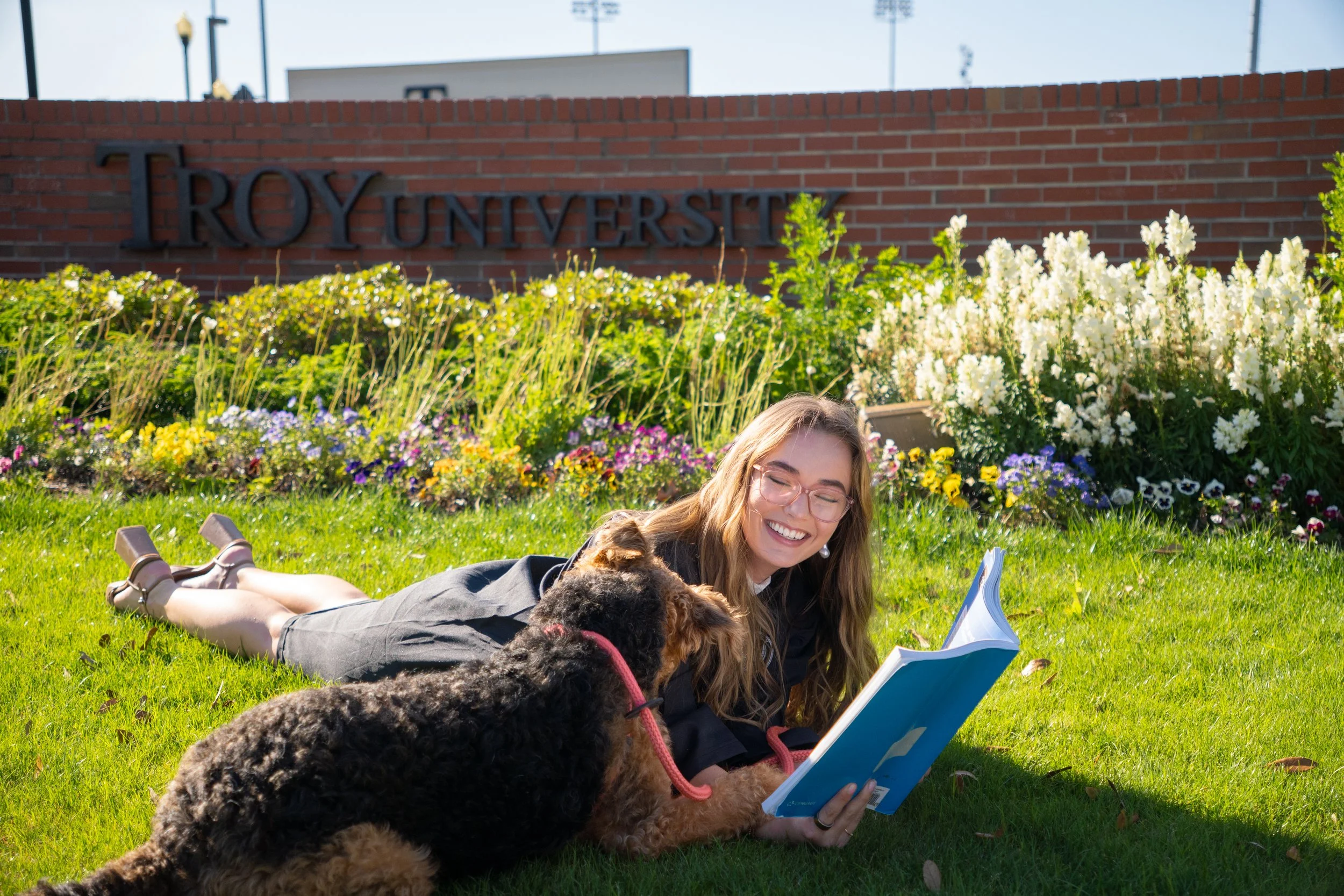 A woman with glasses lies on the grass reading a book, surrounded by two dogs, in front of a flower bed and a brick wall with a university sign that reads 'Troy University'.