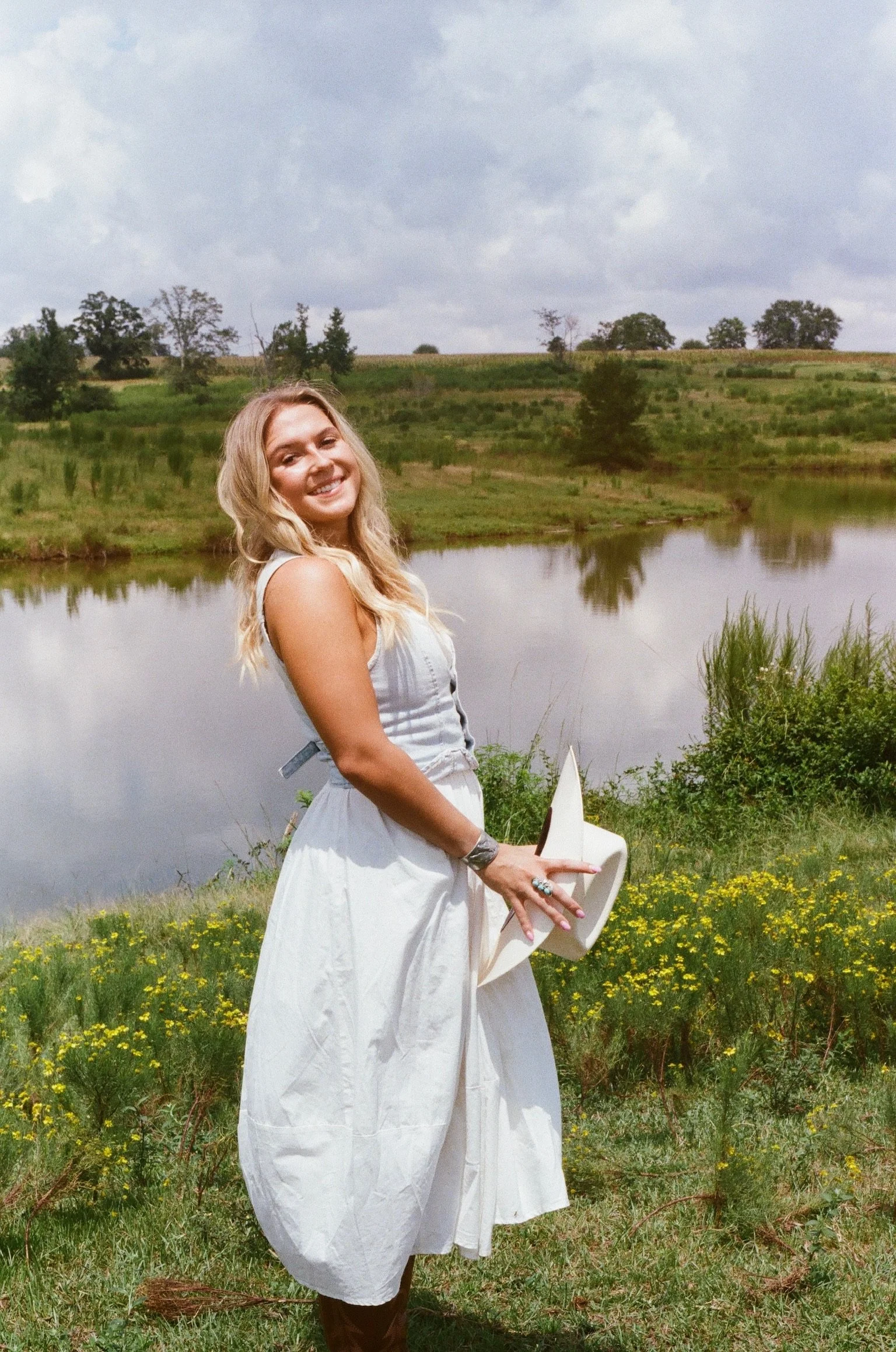 A woman with blonde hair smiling and holding a white wide-brimmed hat, standing in a field of yellow flowers near a body of water with rolling hills and trees in the background.