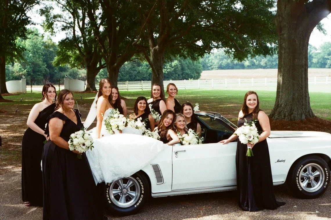 Group of women in black dresses with bouquets posing around a white convertible car in a park with large trees.
