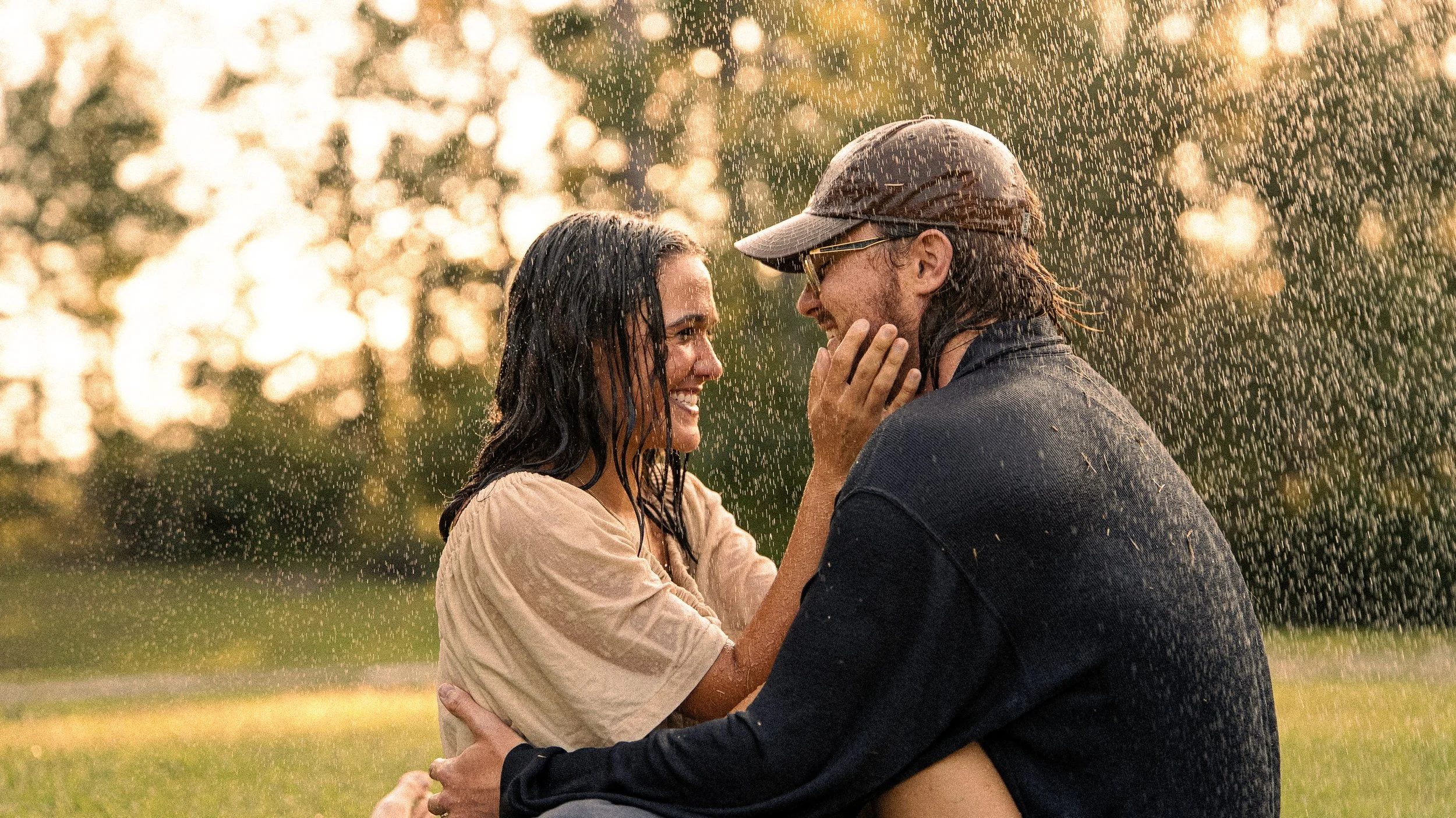 A couple is standing outside in the rain, smiling and looking at each other. The woman has wet hair and wears a light-colored shirt, while the man wears a cap, glasses, and a dark shirt. The background shows sunlight filtering through trees.