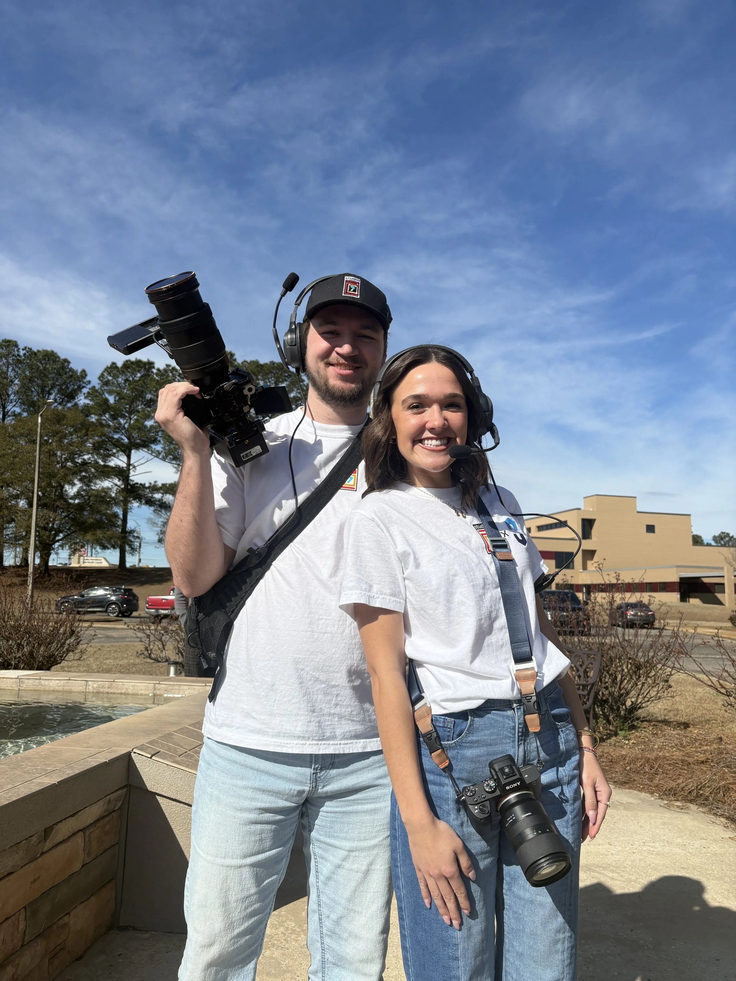 Two young adults standing outdoors with cameras and headsets, smiling, against a backdrop of a blue sky with some clouds, trees, and a building.