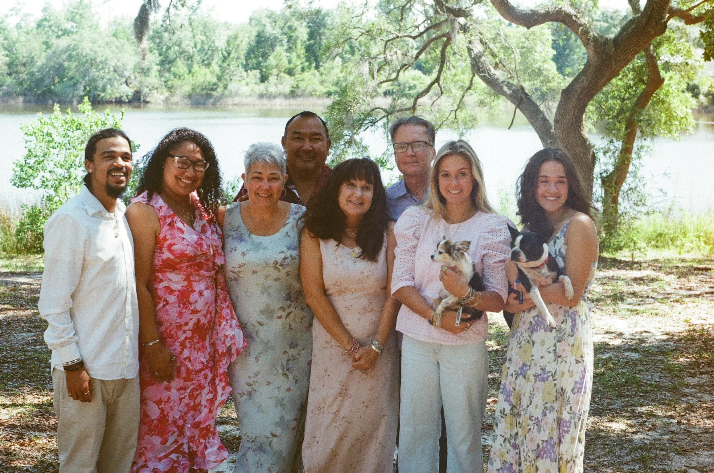 A group of nine people, including women and men, posing outdoors near a lake with green trees and a large branch overhead. Two women are holding small dogs.