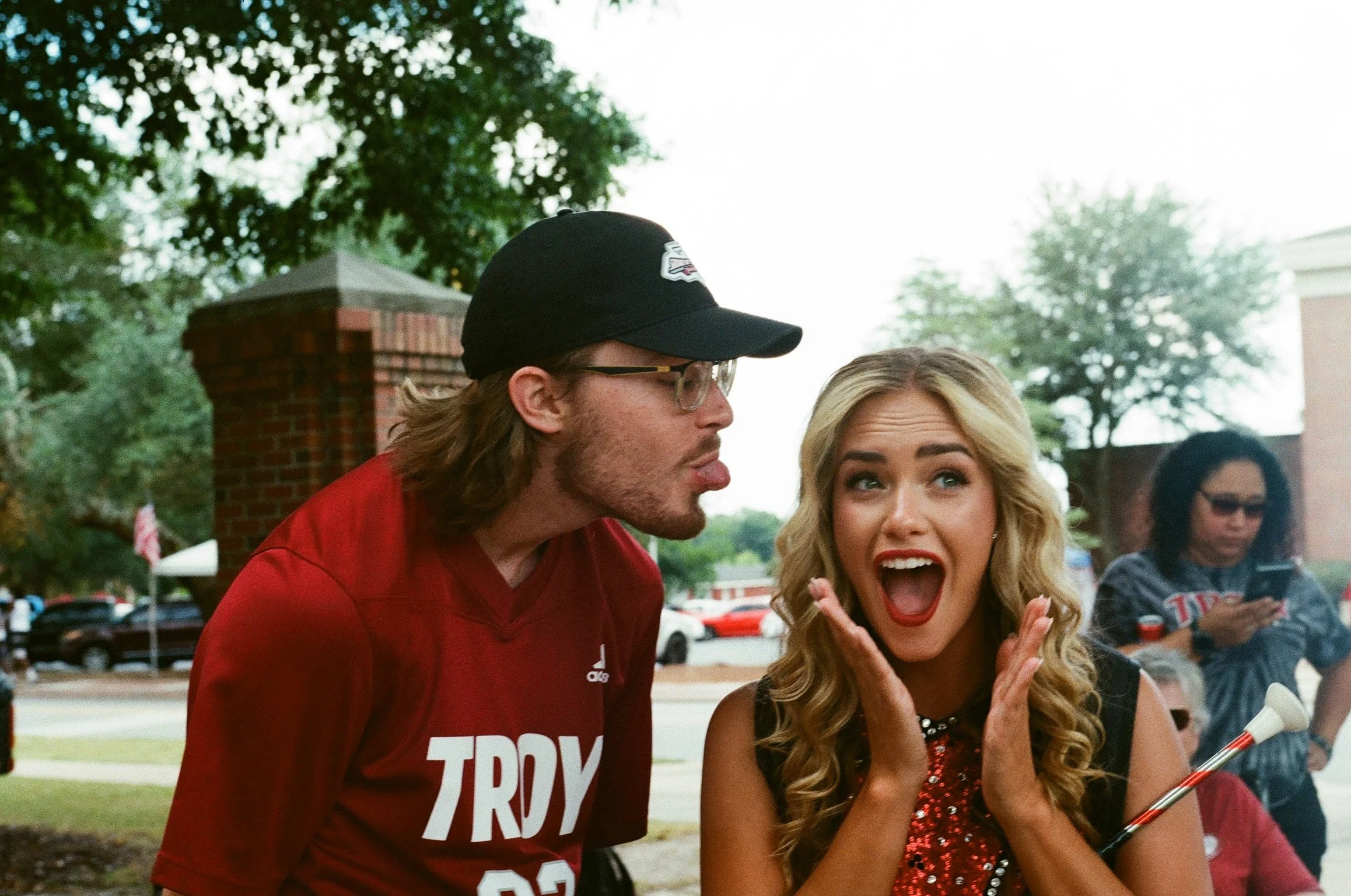 Two people are in foreground of a group gathering outdoors. The man on the left is wearing a black baseball cap, glasses, and a red shirt with white text, and has shoulder-length hair. The woman on the right has long blonde hair and is wearing a sequ