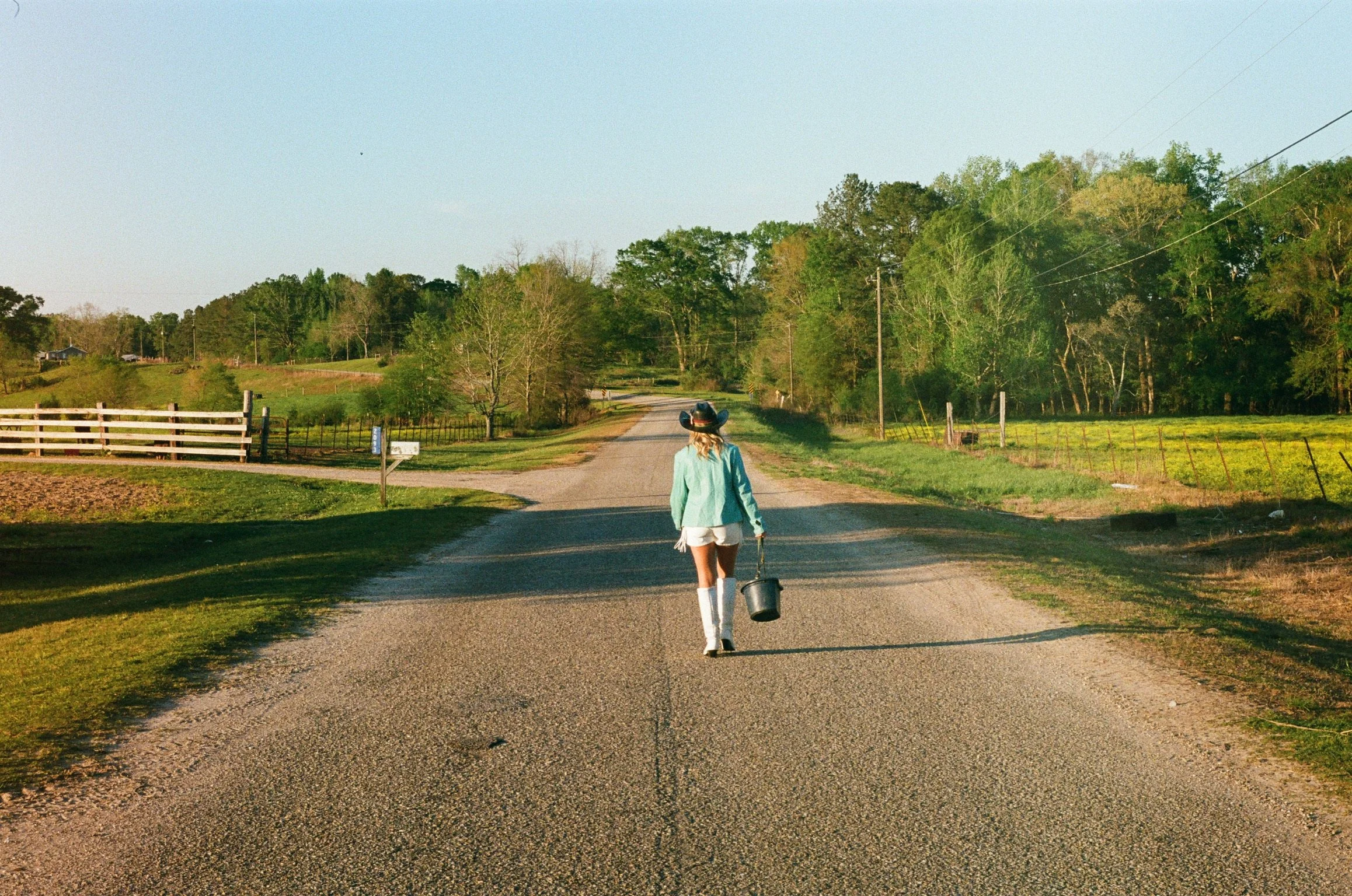 A woman dressed in cowboy boots, a teal jacket, and shorts walking away on a gravel road in a rural area, carrying a black bucket, surrounded by green fields and trees with a clear sky overhead.
