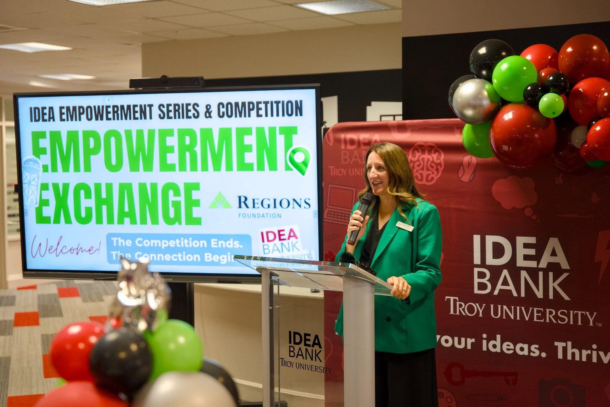 A woman with long brown hair, wearing a green blazer, is speaking into a microphone at a podium during an event. Behind her, there is a large screen displaying the words ‘Idea Empowerment Series & Competition’ and ‘Empowerment’ in bright green text, 