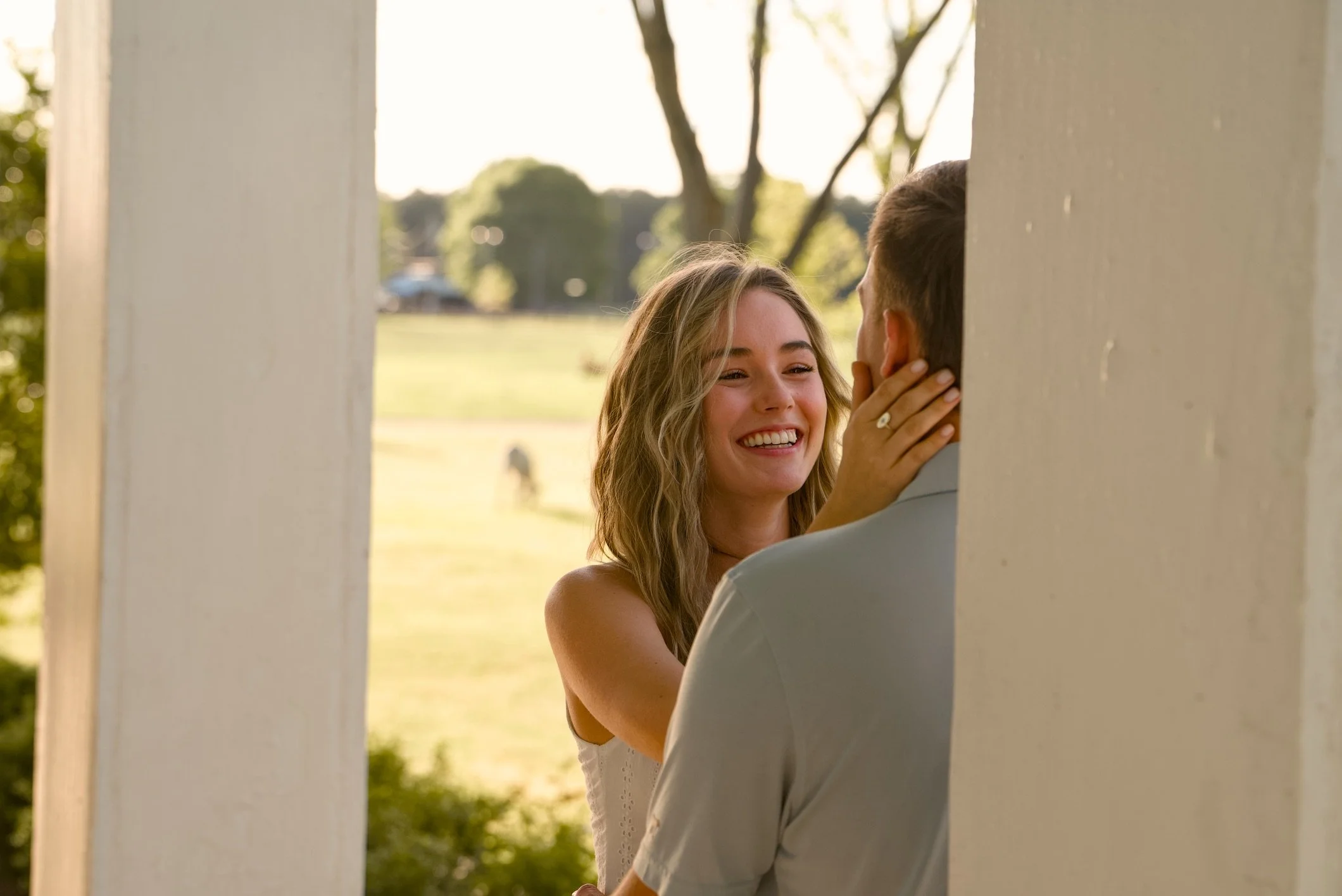A smiling woman and a man are close to each other, touching faces and holding hands outside, framed by white columns.