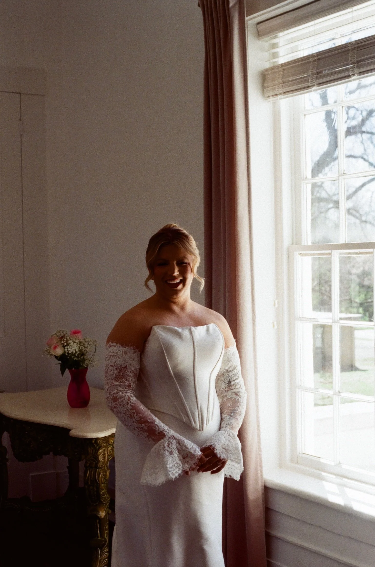 A smiling woman wearing a white wedding dress with lace sleeves, standing near a window with curtains, holding her hands together in front of her.