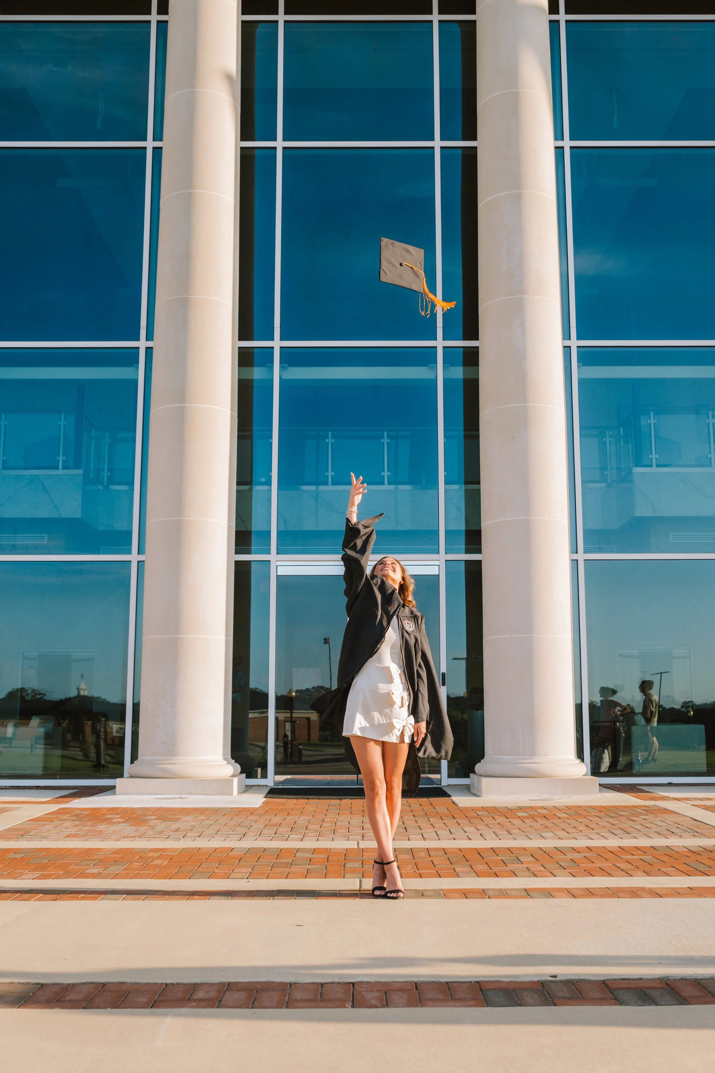 A woman in a graduation gown and shorts standing outside in front of a modern glass building, throwing her graduation cap into the air.