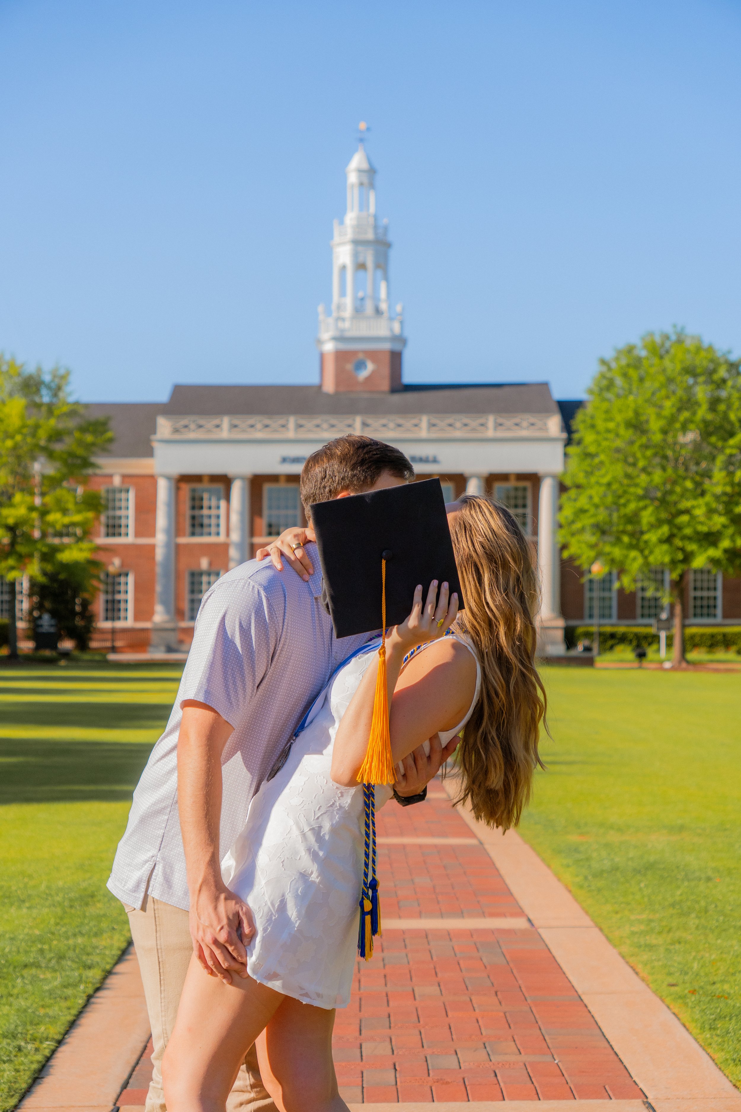 A couple embracing during graduation on a college campus with a historic building and a steeple in the background.