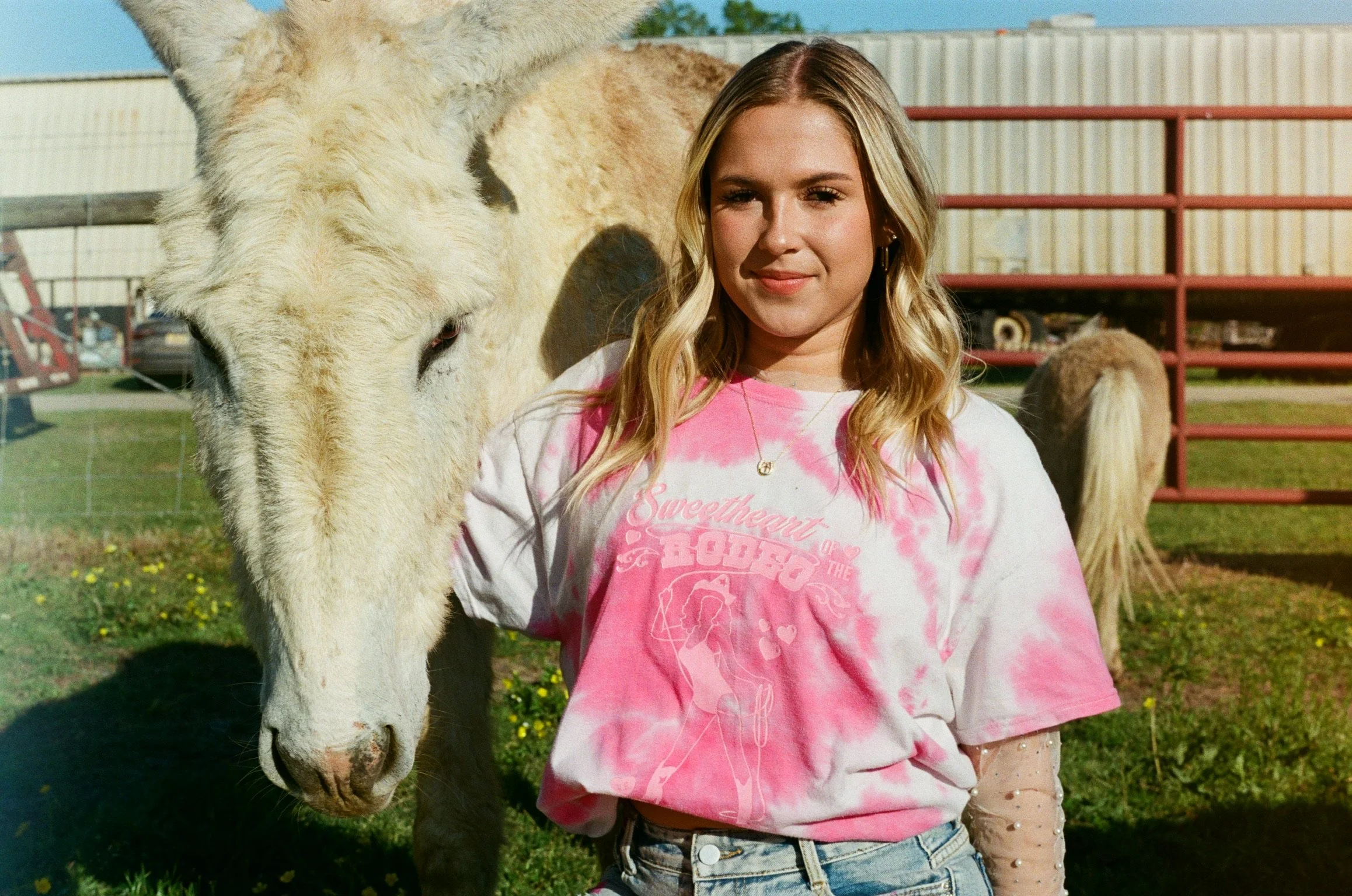 A young woman standing next to a white horse outdoors on a sunny day, with a fence and industrial buildings in the background.