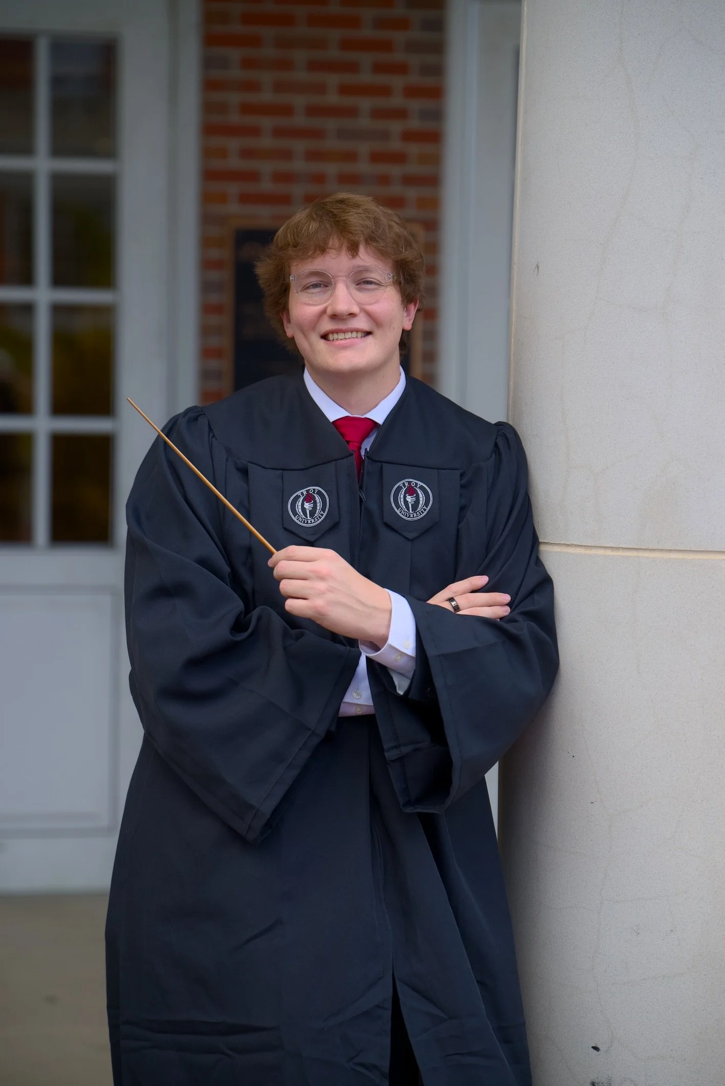 A young man smiling in graduation attire, standing outdoors with arms crossed, holding a diploma, wearing glasses, and a black gown with a red tie, with a brick building and window in the background.
