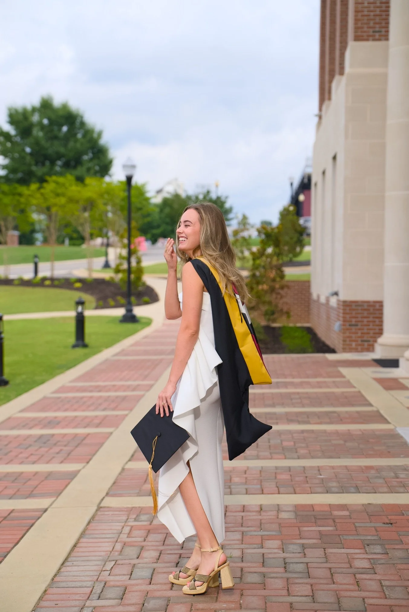 A young woman in a white dress and high heels smiling while holding a graduation cap and wearing a black graduation gown with yellow accents, standing outdoors on a brick pathway with trees and lampposts in the background.