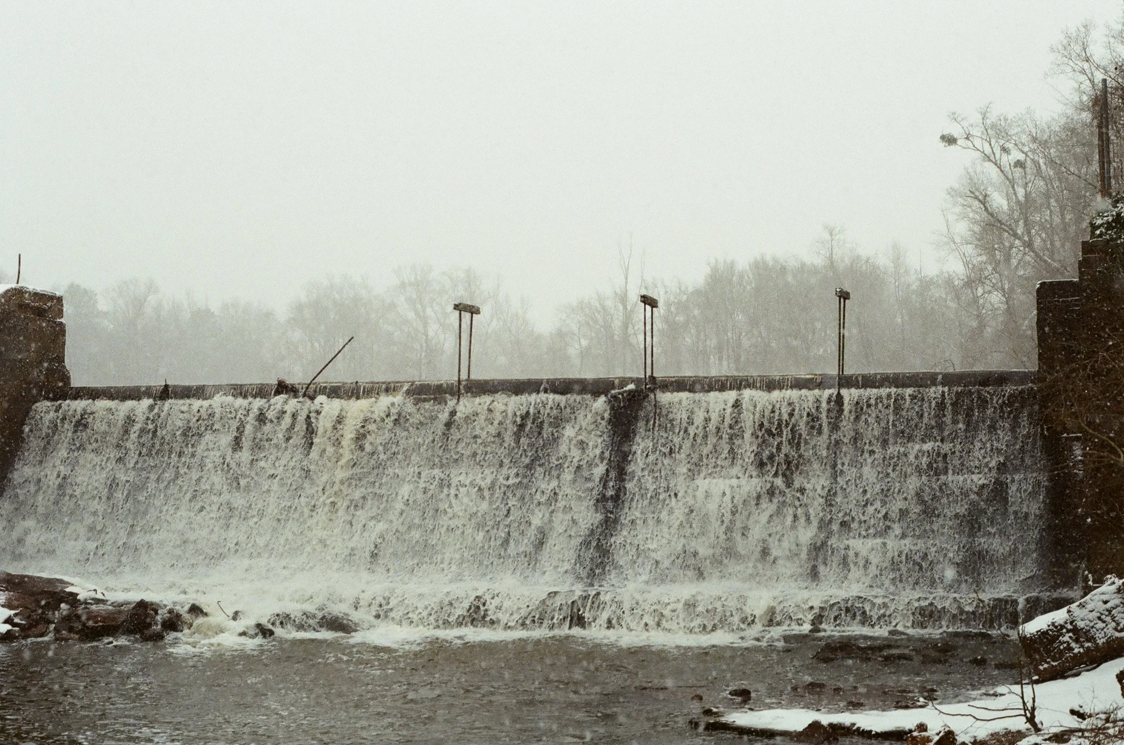 Water flowing over a dam or waterfall with trees and overcast sky in the background, snow on the ground.
