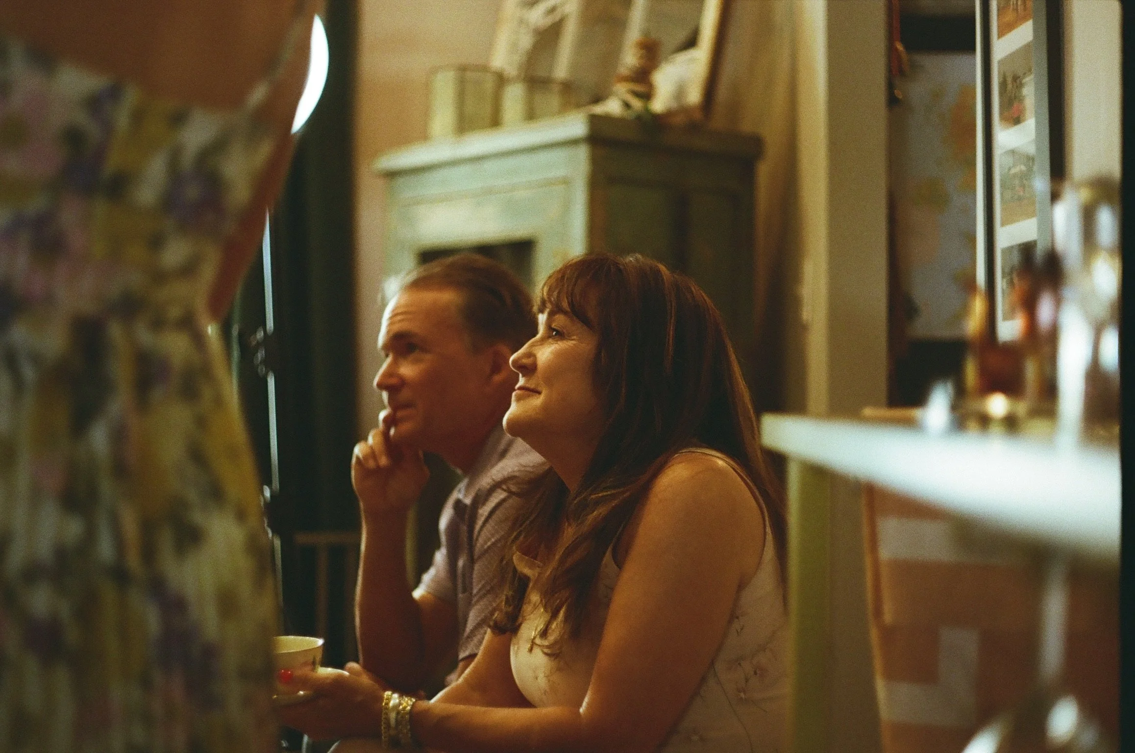 Two people, a man and a woman, sit at a table in a cozy indoor setting. They are smiling and appear to be enjoying each other's company. The woman has dark red hair and is wearing a light-colored top, while the man has light brown hair and is wearing