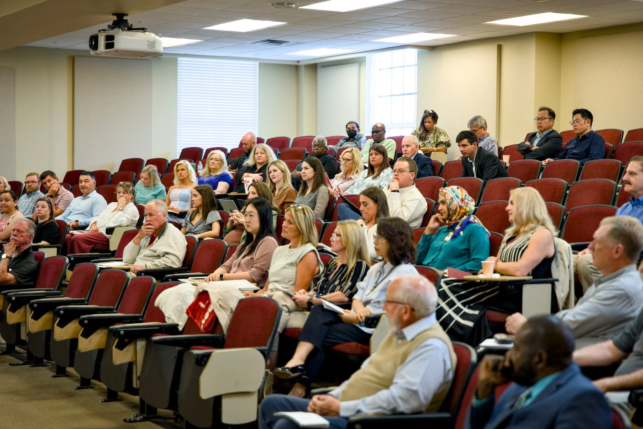 Audience of diverse adults sitting in a lecture hall during a presentation, some taking notes, some listening attentively.