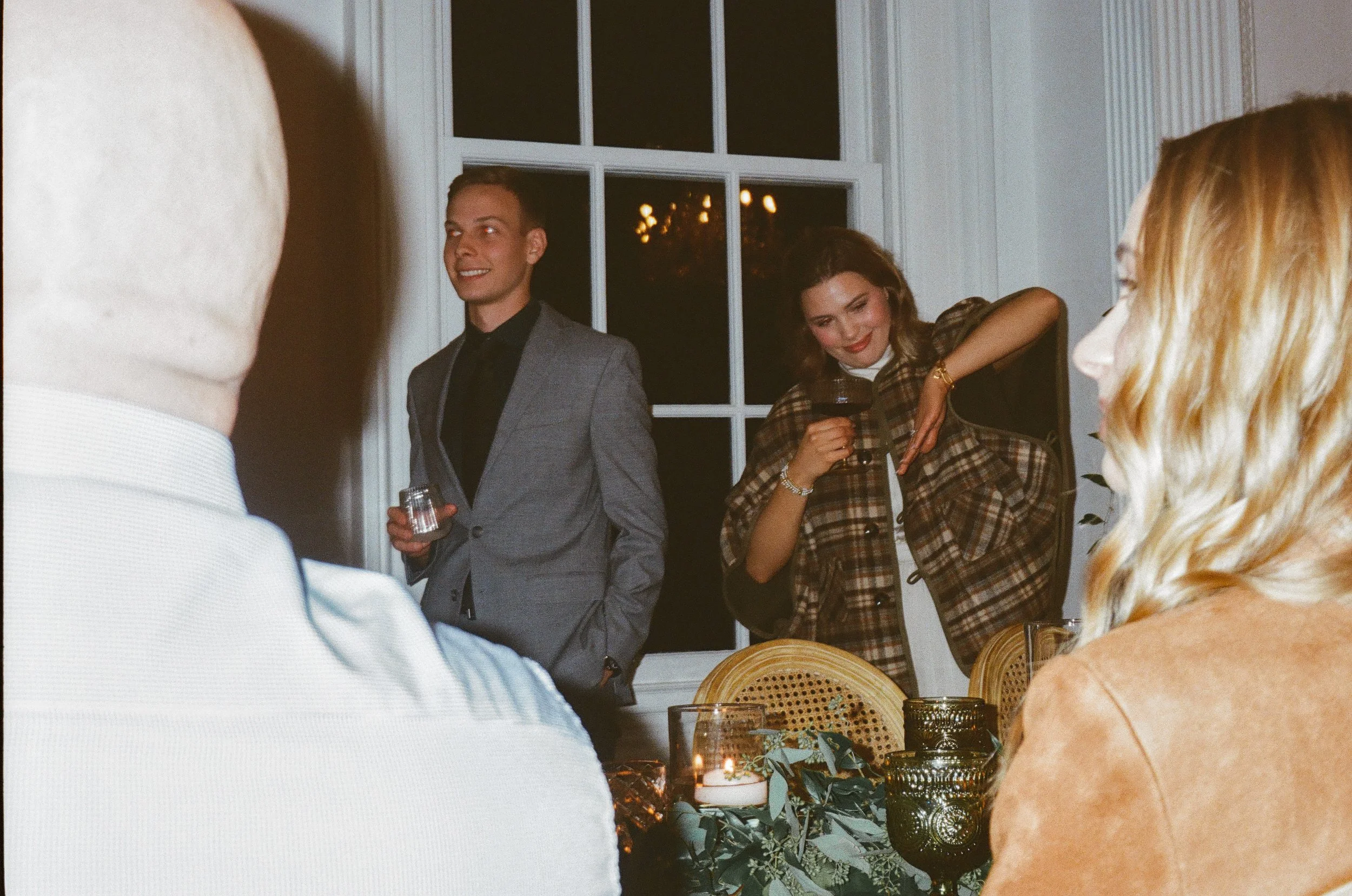 People gathered at a dinner party, with a man in a gray suit holding a glass and a woman in plaid smiling, while others are seated around a table decorated with candles and greenery.