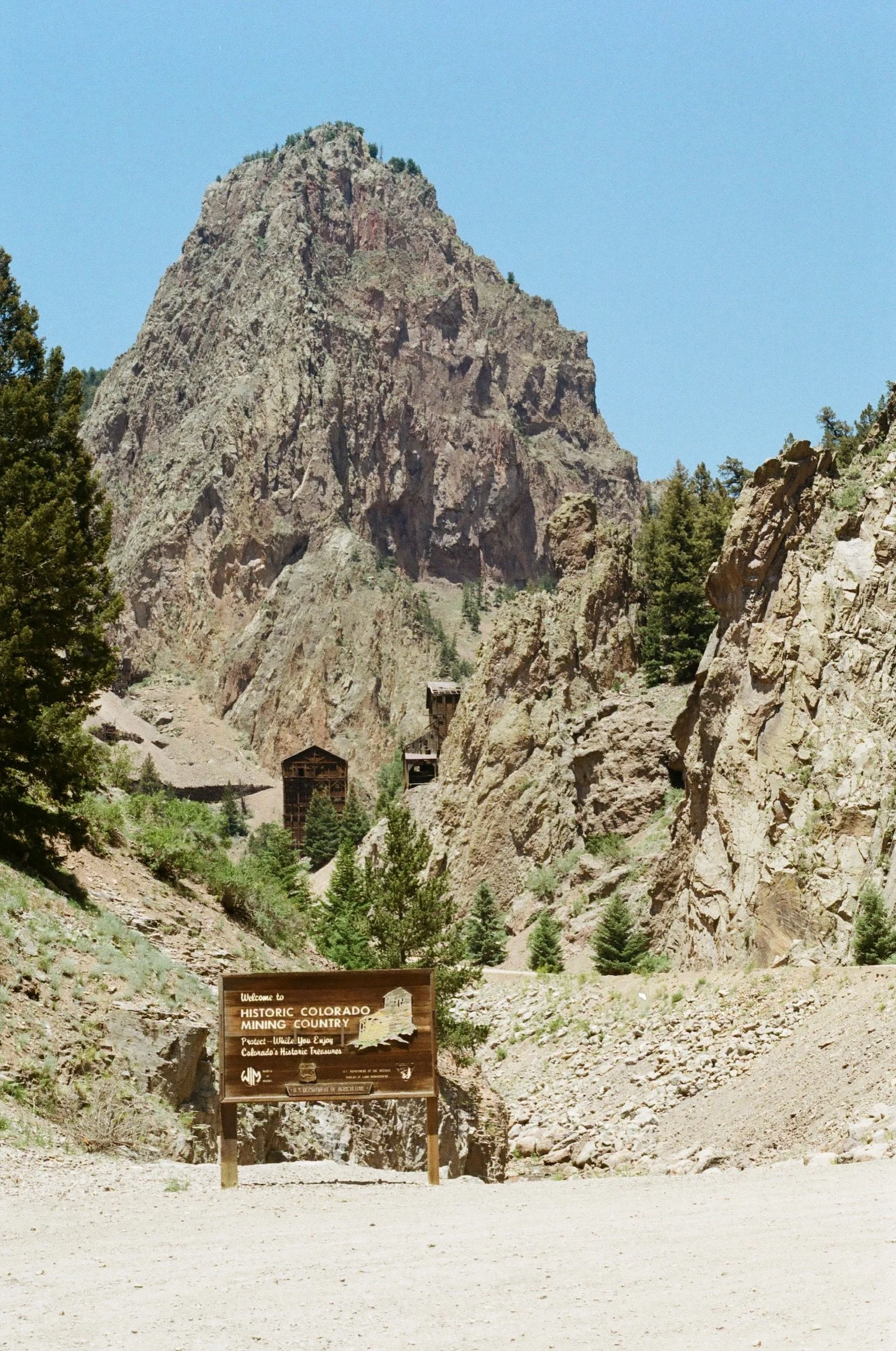 Large rocky mountain with green trees at its base and a blue sky. A wooden sign at the bottom reads 'Welcome to Historic Colorado Mining Country'.