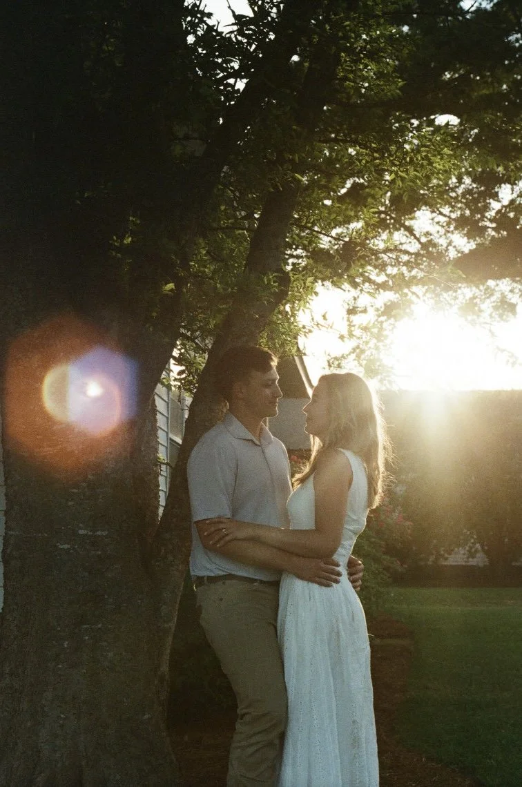 A couple standing close together outdoors at sunset, with the man leaning against a tree and the woman facing him, both gazing into each other's eyes.