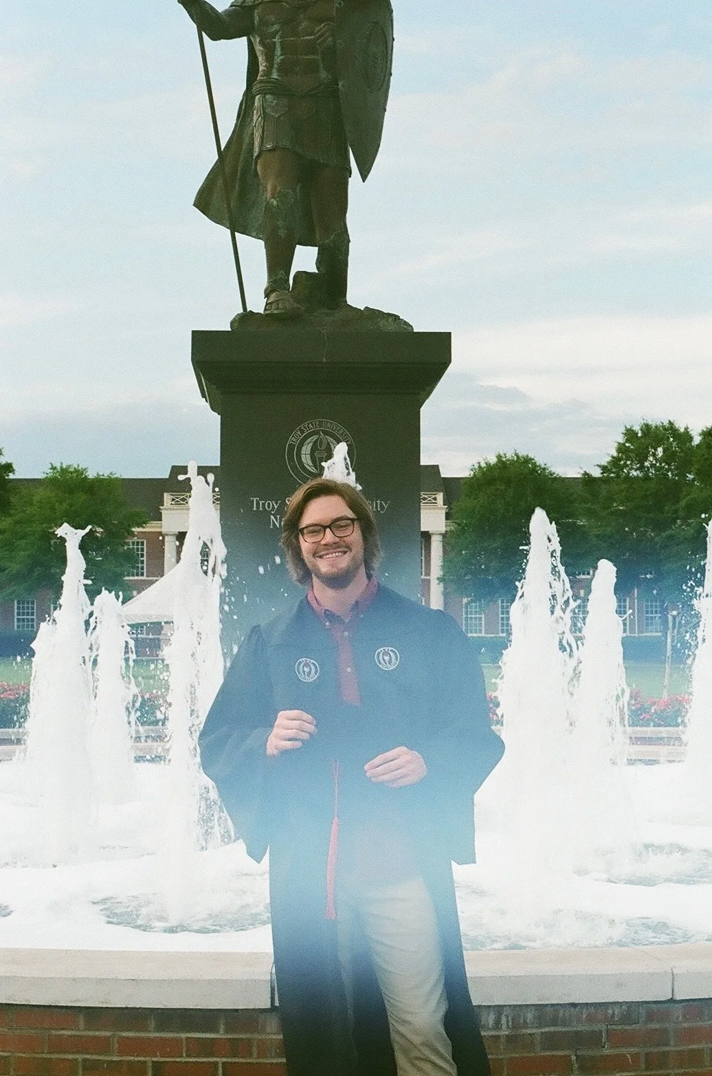 A young man standing in front of a fountain with water jets, smiling and holding a diploma or rolled document in his hand. Behind him, there is a large statue of a historical figure in front of a university building, with trees and a cloudy sky in th