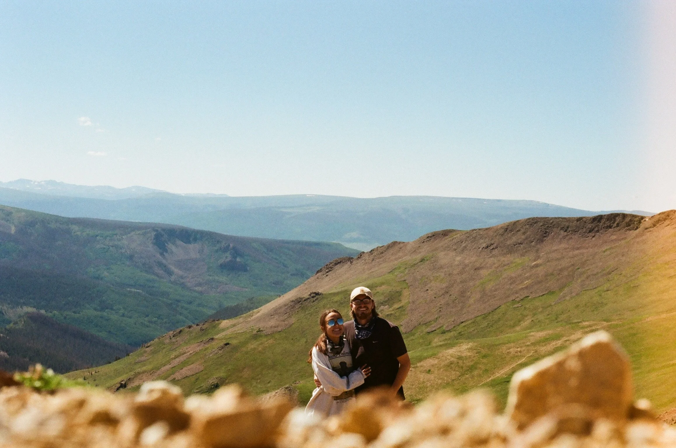Two people, a man and a woman, standing in a scenic mountainous landscape with rolling green hills and distant mountains under a bright blue sky. The woman is embracing the man, and both are smiling.