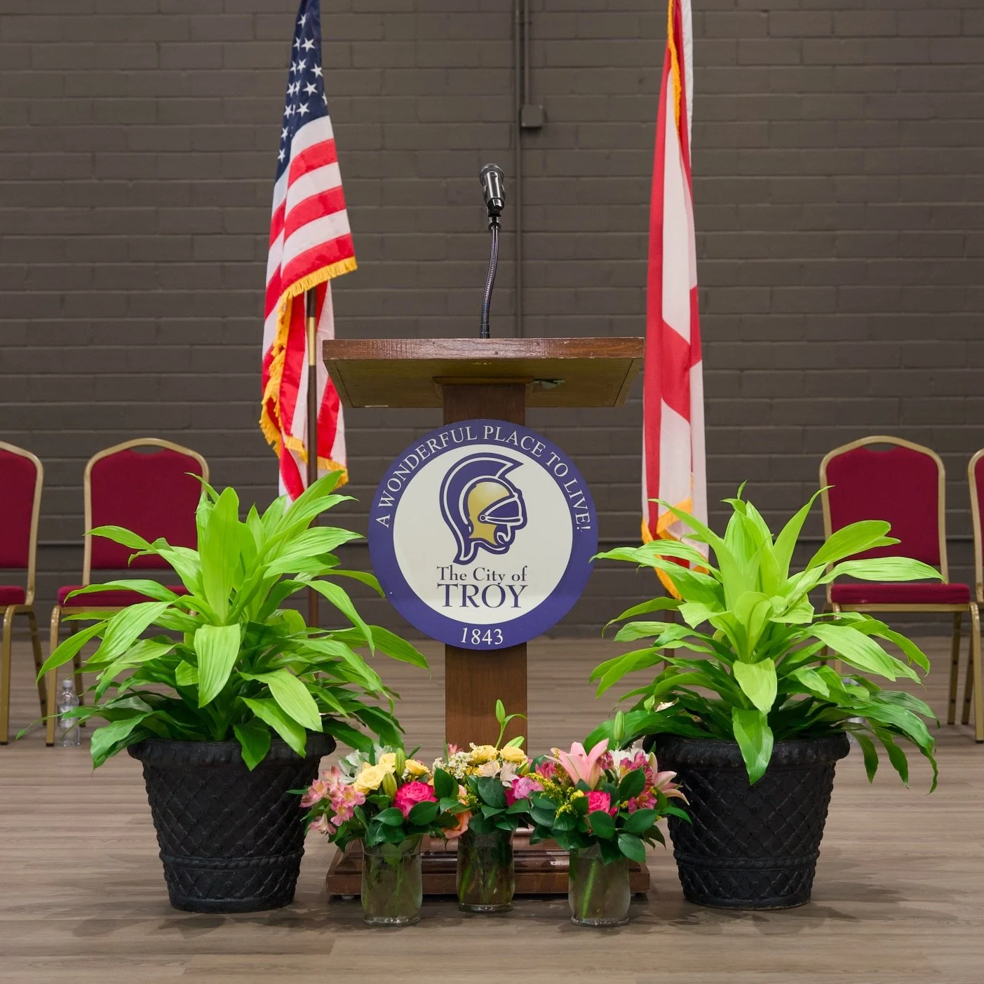 A wooden podium with a microphone, decorated with the City of Troy emblem, flanked by American and state flags. There are potted green plants and colorful flowers in front of the podium, with chairs in the background against a dark brick wall.