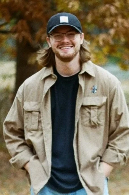 A young man with long hair, glasses, and a beard, smiling outdoors in autumn, wearing a black cap, tan jacket, and black shirt.