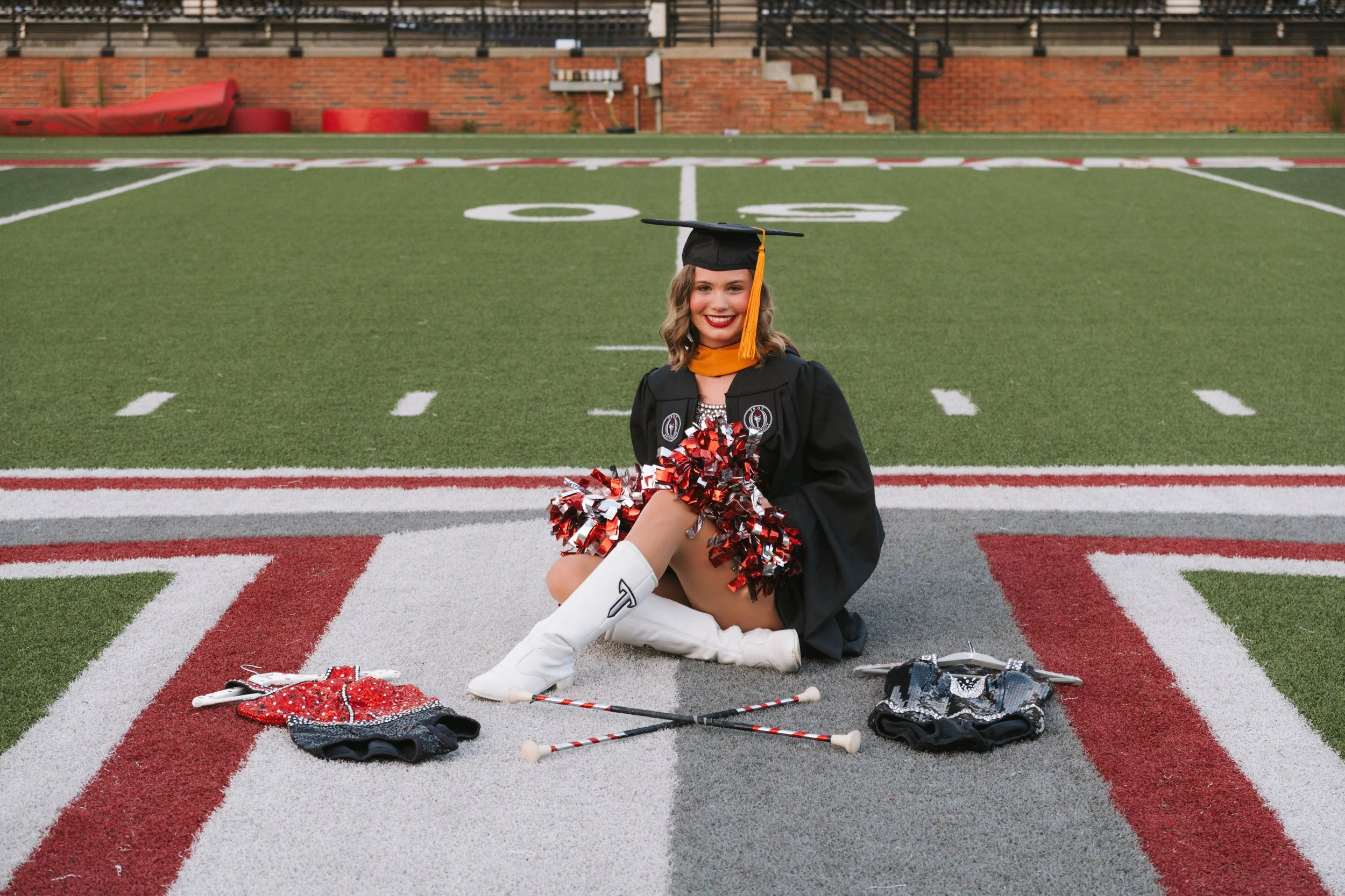 A young woman in a graduation cap and gown sitting on a football field, smiling, holding pom-poms, with cheerleading accessories and a golf club on the ground nearby.