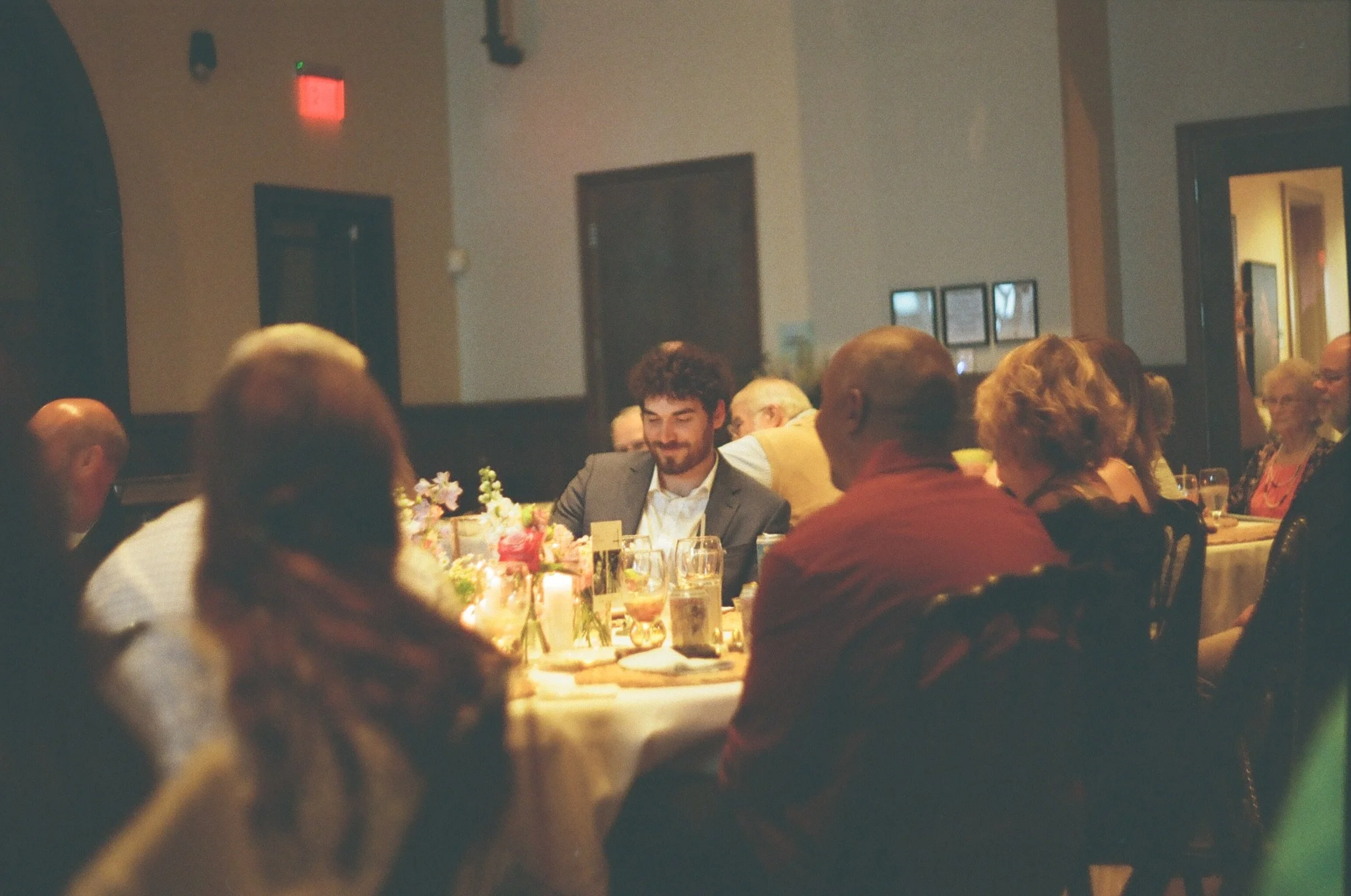 A gathering of people seated around a table at a formal event, with some floral centerpieces and drinks on the table. The background shows a beige wall with framed pictures and an open door.