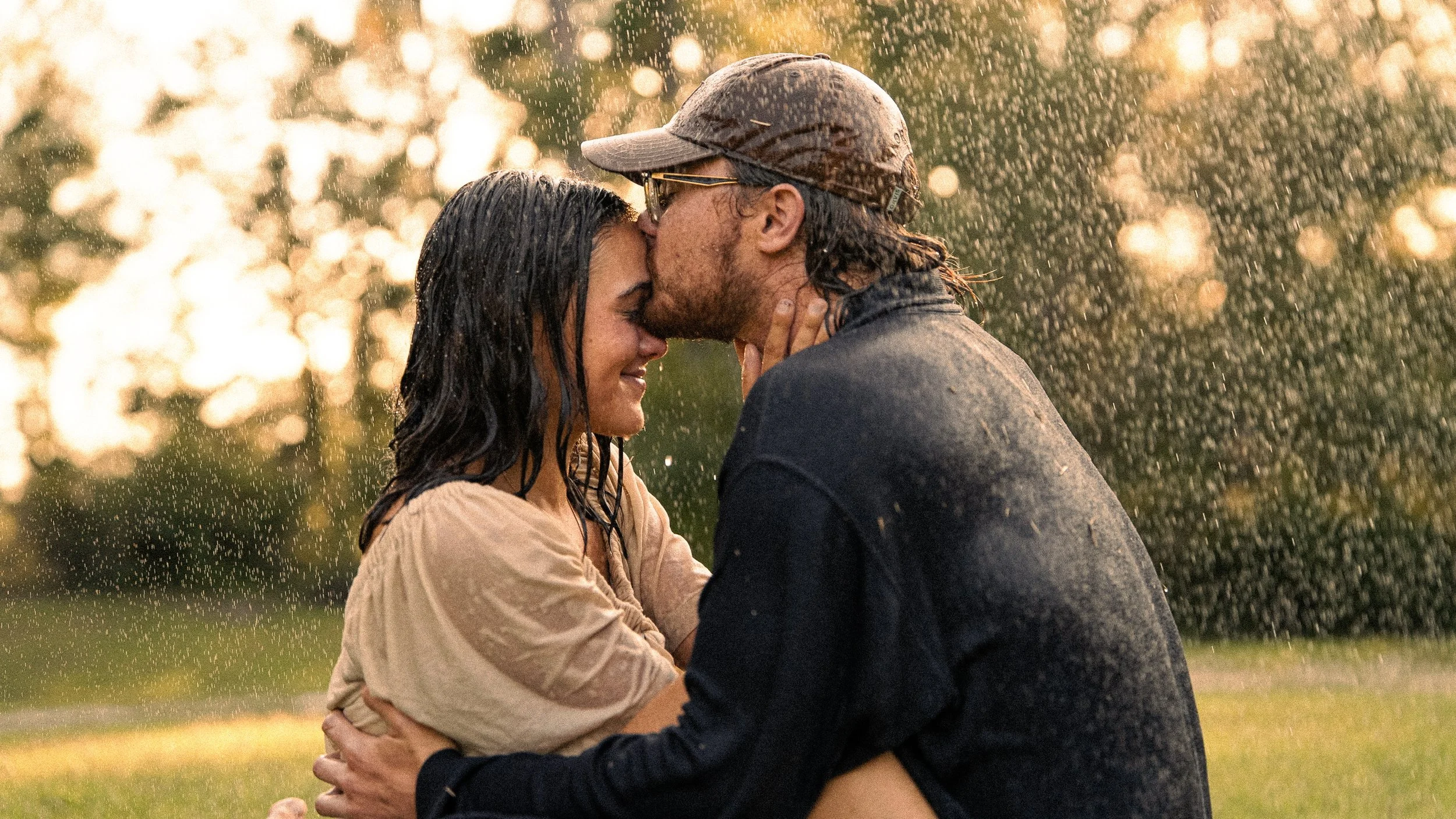 A man and a woman kissing outdoors in the rain, with trees in the background and sunlight filtering through.