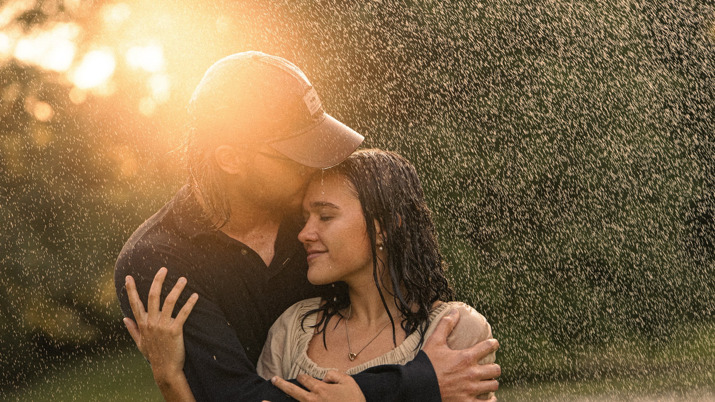 Two women embracing in the rain during sunset, with water droplets illuminated by the sunlight.