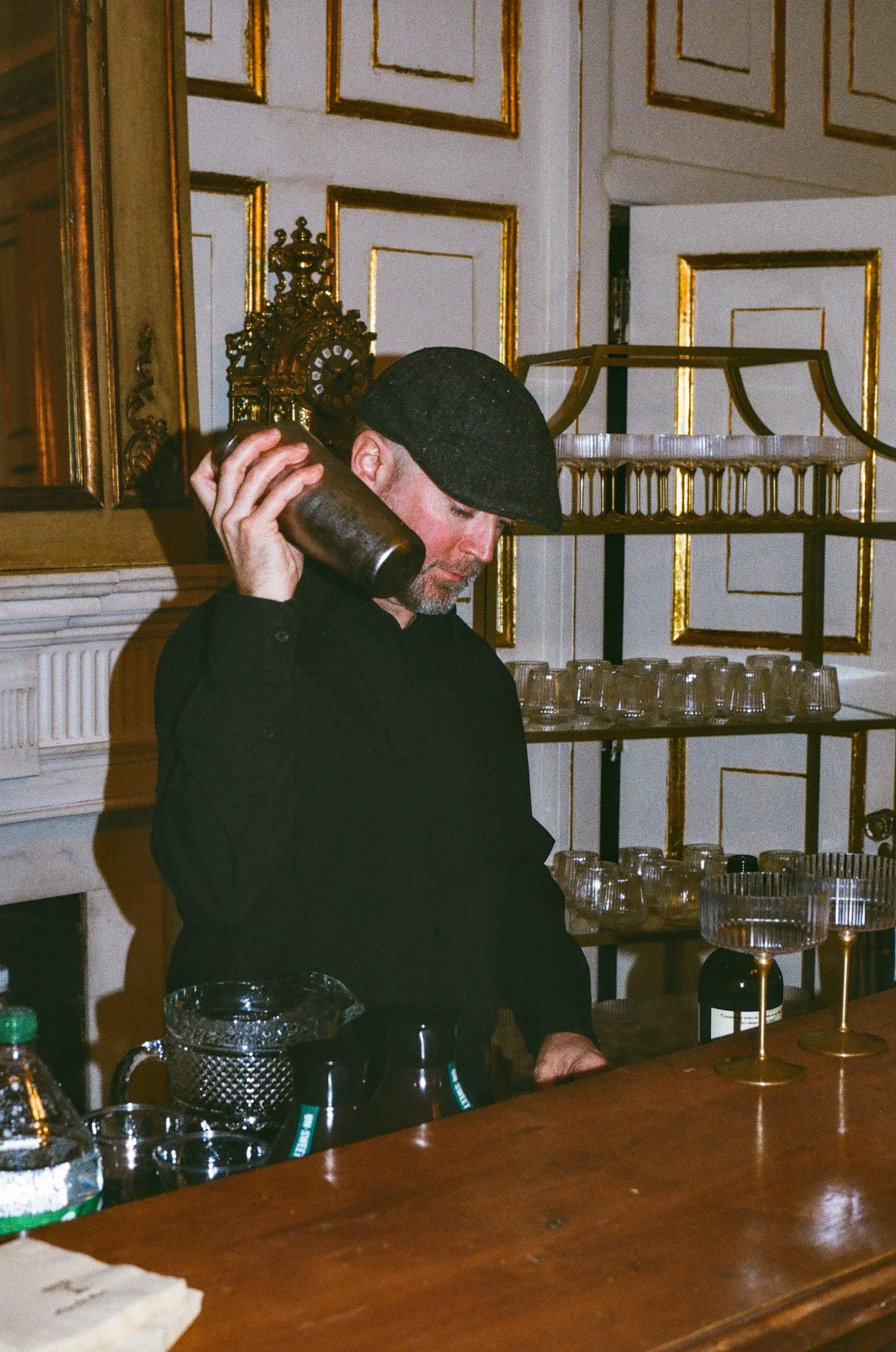 A man wearing a black cap and black shirt behind a bar, pouring a drink. The bar is decorated with glassware and bottles, with an ornate gold and white wall in the background.