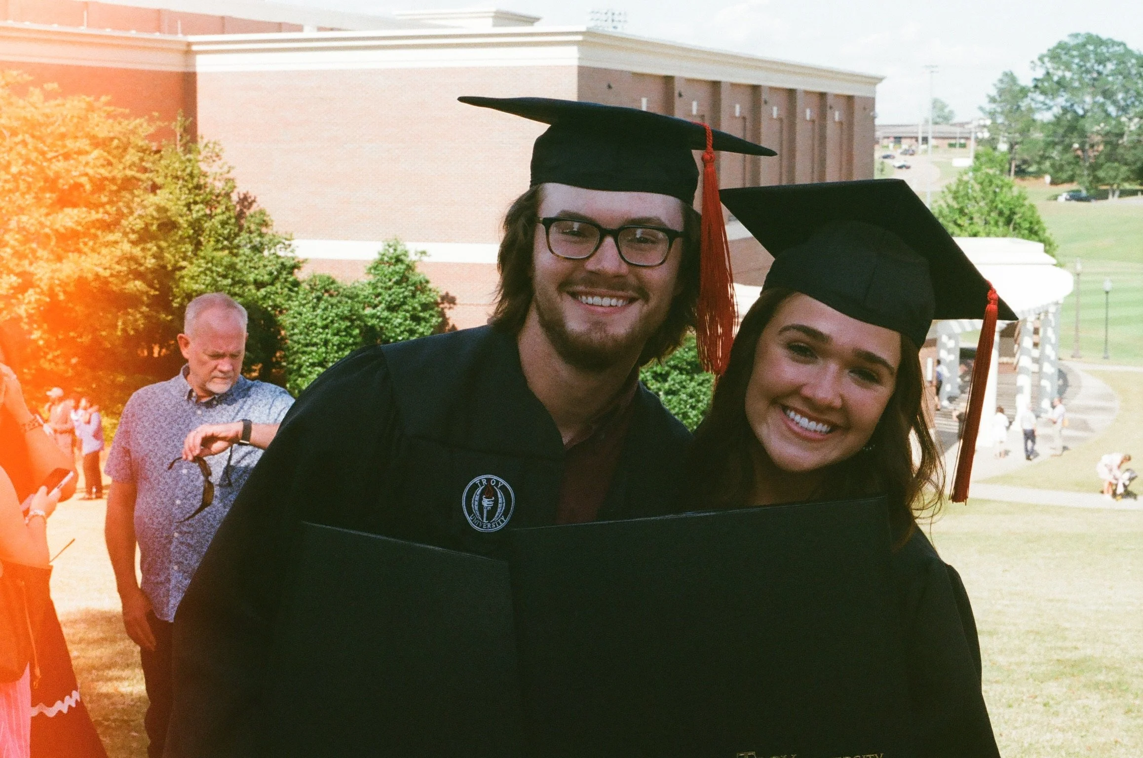 Two graduates in black caps and gowns smiling, attending a graduation ceremony outdoors on a sunny day, with other people and a large brick building in the background.