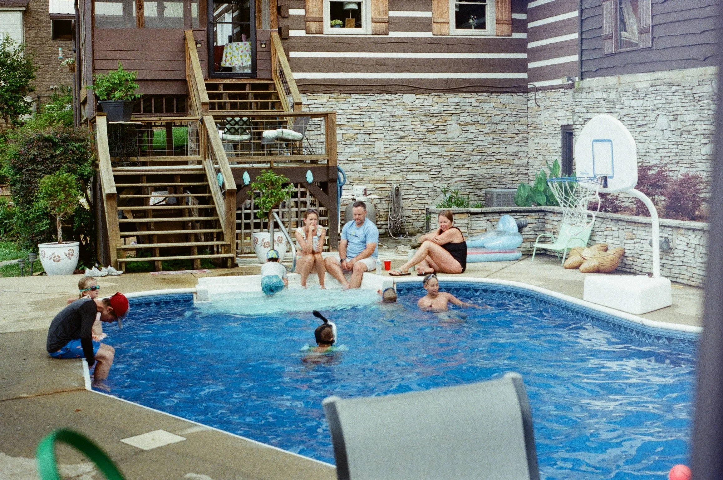 A backyard pool scene with children and adults, with a wooden deck, stone wall, and a basketball hoop in the background. People are swimming, sitting, and playing around the pool.