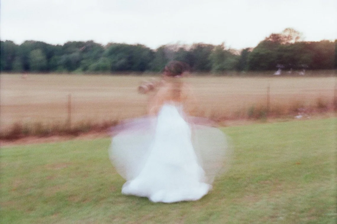 A person wearing a white dress, spinning in a grassy field with trees and a fence in the background, captured in motion blur.