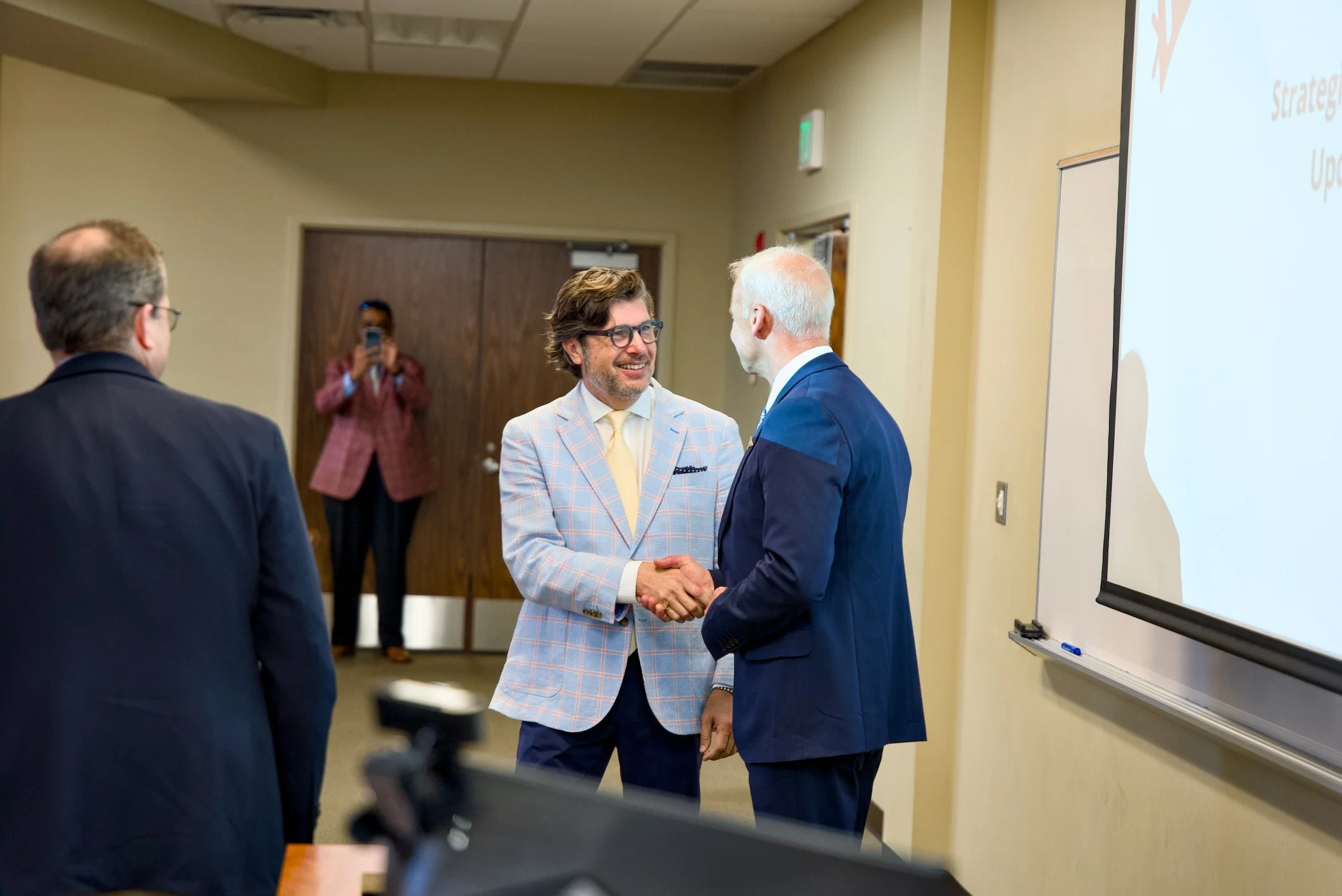 Two men shaking hands at a professional event, with a woman taking a photo in the background and a whiteboard screen on the right.