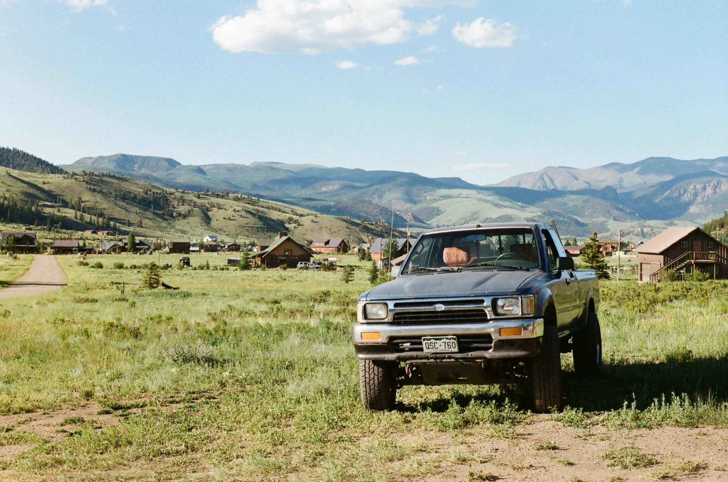A black pickup truck parked on a grassy field in a rural area with houses, trees, and mountains in the background, under a blue sky with some white clouds.