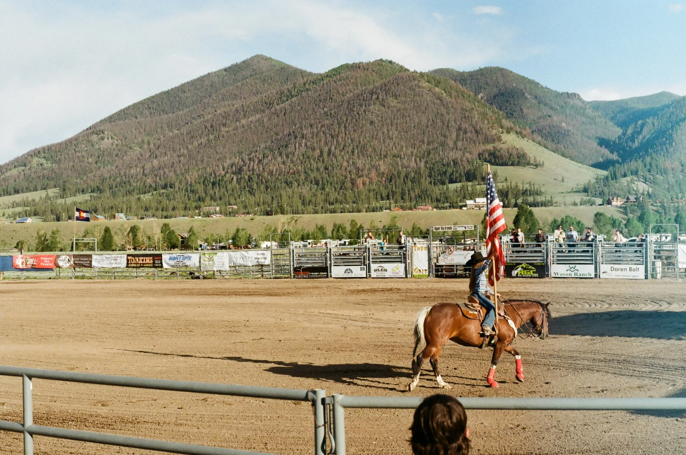 A rodeo arena with a rider on a horse holding an American flag, and mountains in the background.