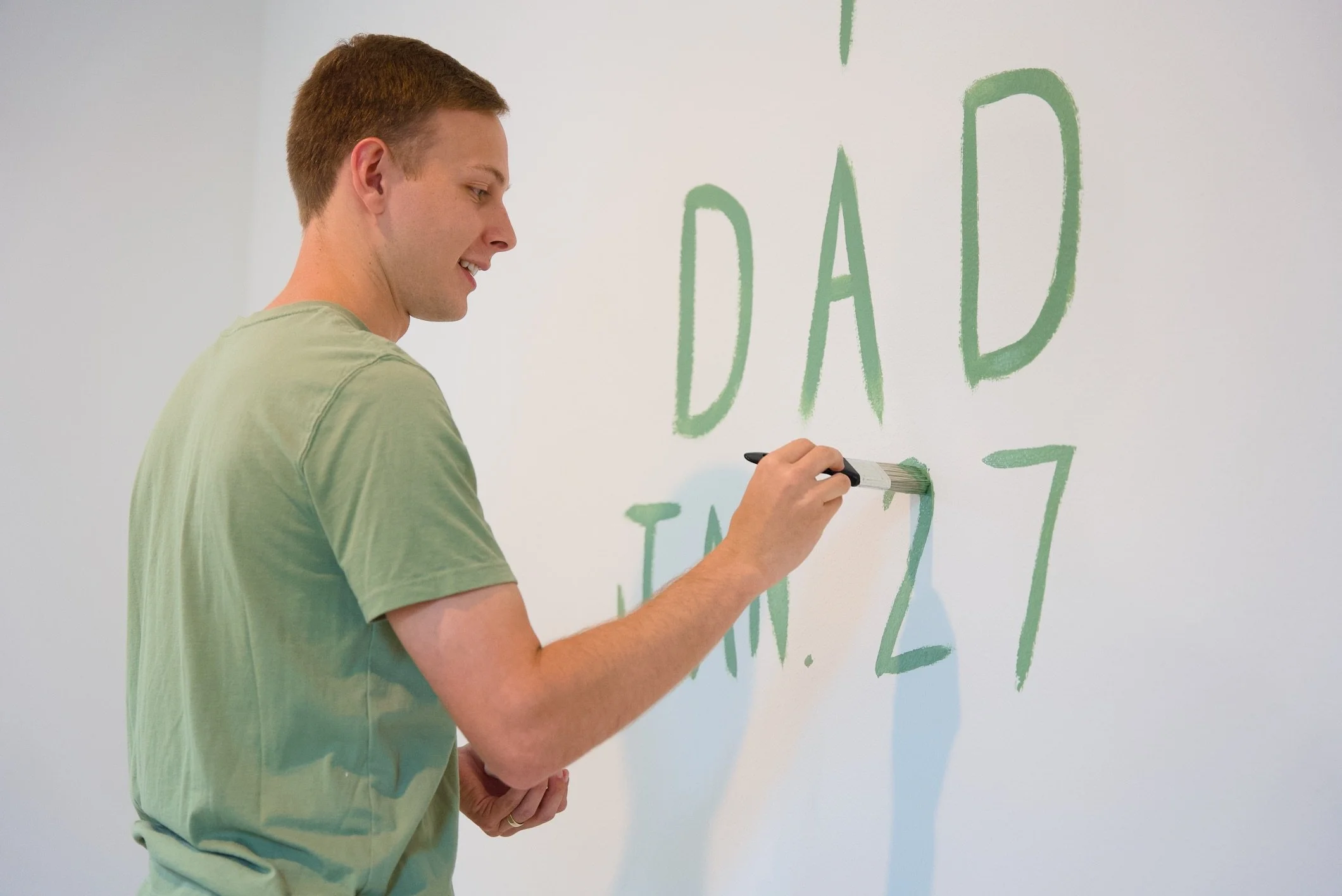 A man in a light green t-shirt writing on a white wall with a green marker. The wall has green text reading 'DAD' and partially visible words.